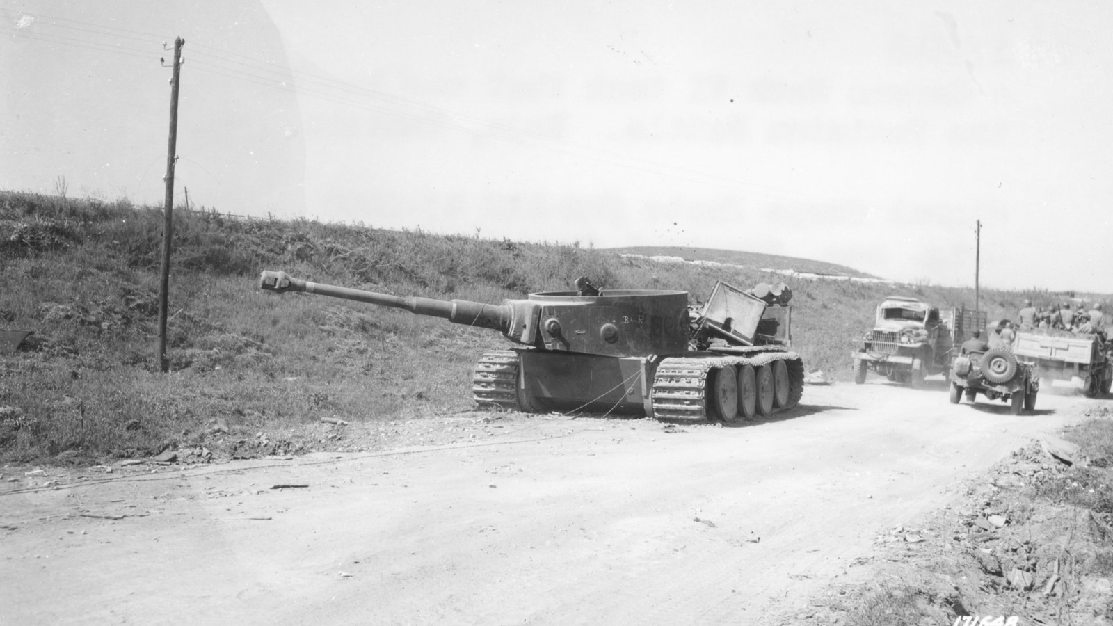 Black and white photograph of a wrecked German Tiger tank at the side of a dirt road with its long gun pointing left, while an Allied truck and jeep drive past on the road behind it.