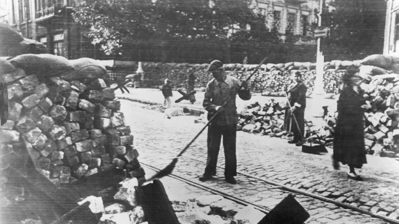 Black and white photograph of a cobbled street in Odessa with tram rails, walls of stacked paving stones and sandbags across the road, a man sweeping in the middle and a woman walking past.