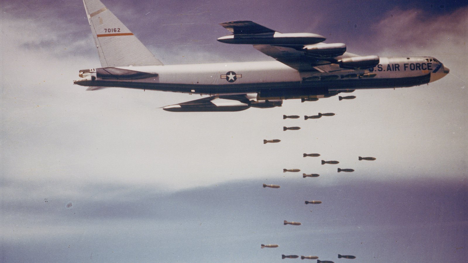 Color photograph of a US Air Force B-52 bomber in flight releasing a long string of bombs against a hazy sky.