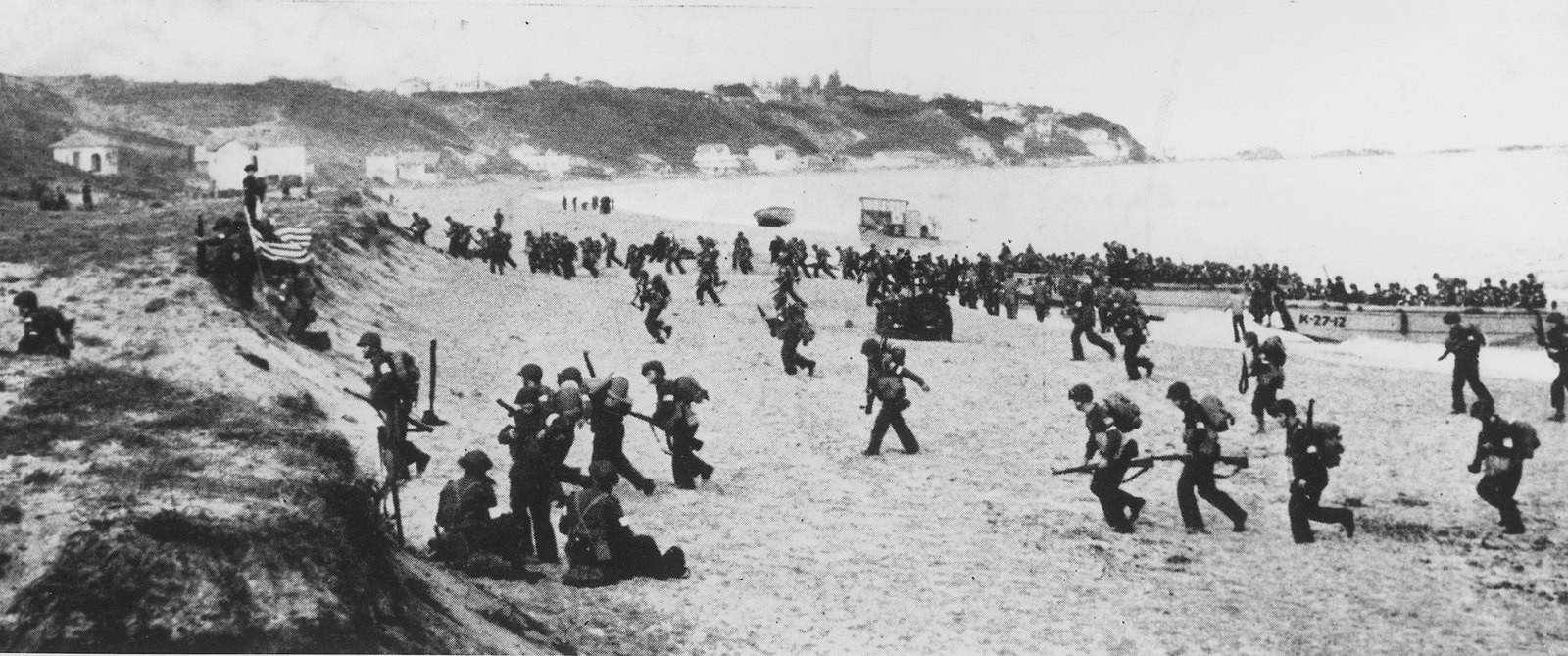 Black and white photograph of helmeted soldiers running up a wide sandy beach from landing craft towards a dune, where one man holds an American flag, with white houses on a hill behind