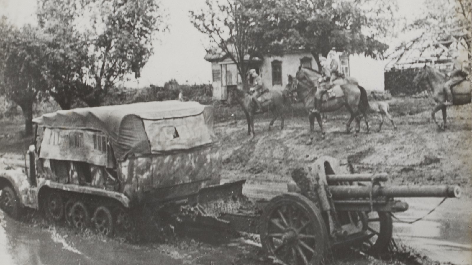 Old black and white photograph of a German half-track with a canvas cover towing a field howitzer through a flooded, muddy village street, with horsemen and a house behind