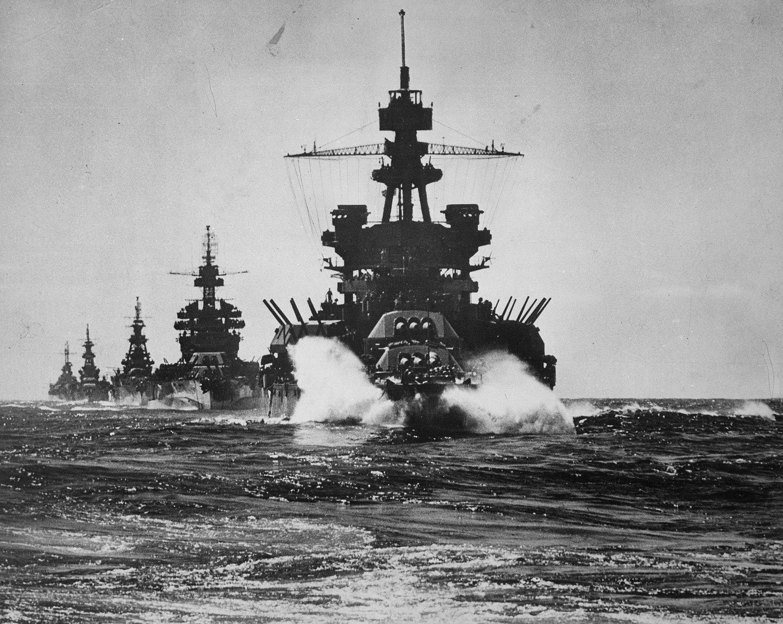 Black and white photograph of a line of warships at sea: a battleship with a tall tripod mast and spray at its bow leads, followed by another battleship and several cruisers.