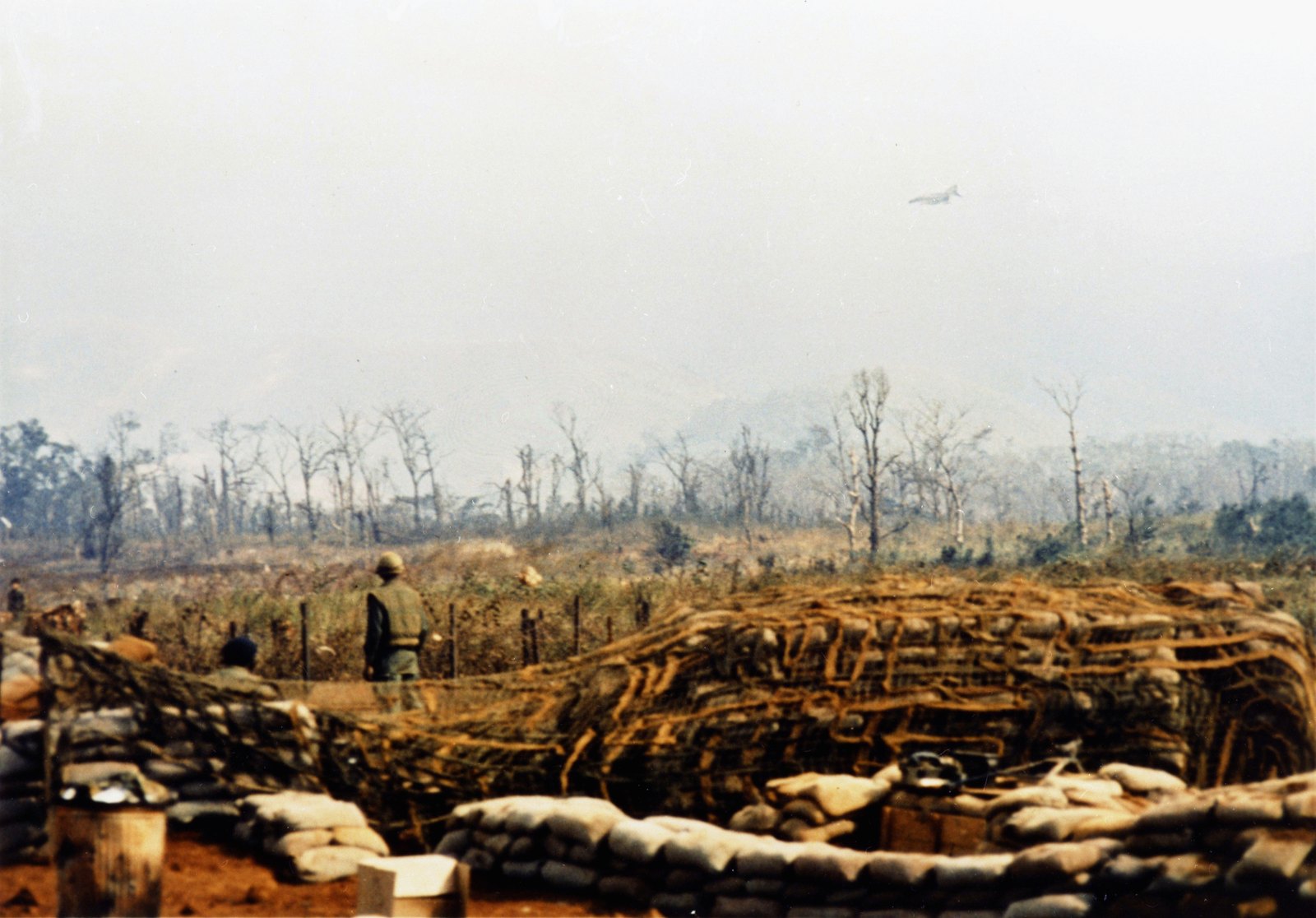 Color photograph of sandbagged and camouflage-netted bunkers at Khe Sanh in the foreground, a Marine standing by the wire, shattered trees behind, and a jet aircraft low in the hazy sky.