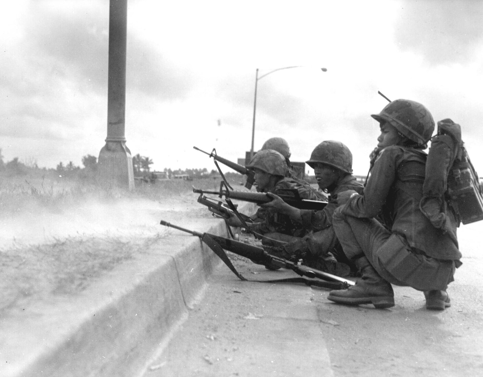 Black and white photograph of four South Vietnamese Rangers in helmets crouching behind a concrete curb on a Saigon roadside, rifles pointed over the edge, one with a radio on his back, a street lamp above them and smoke drifting across the ground.