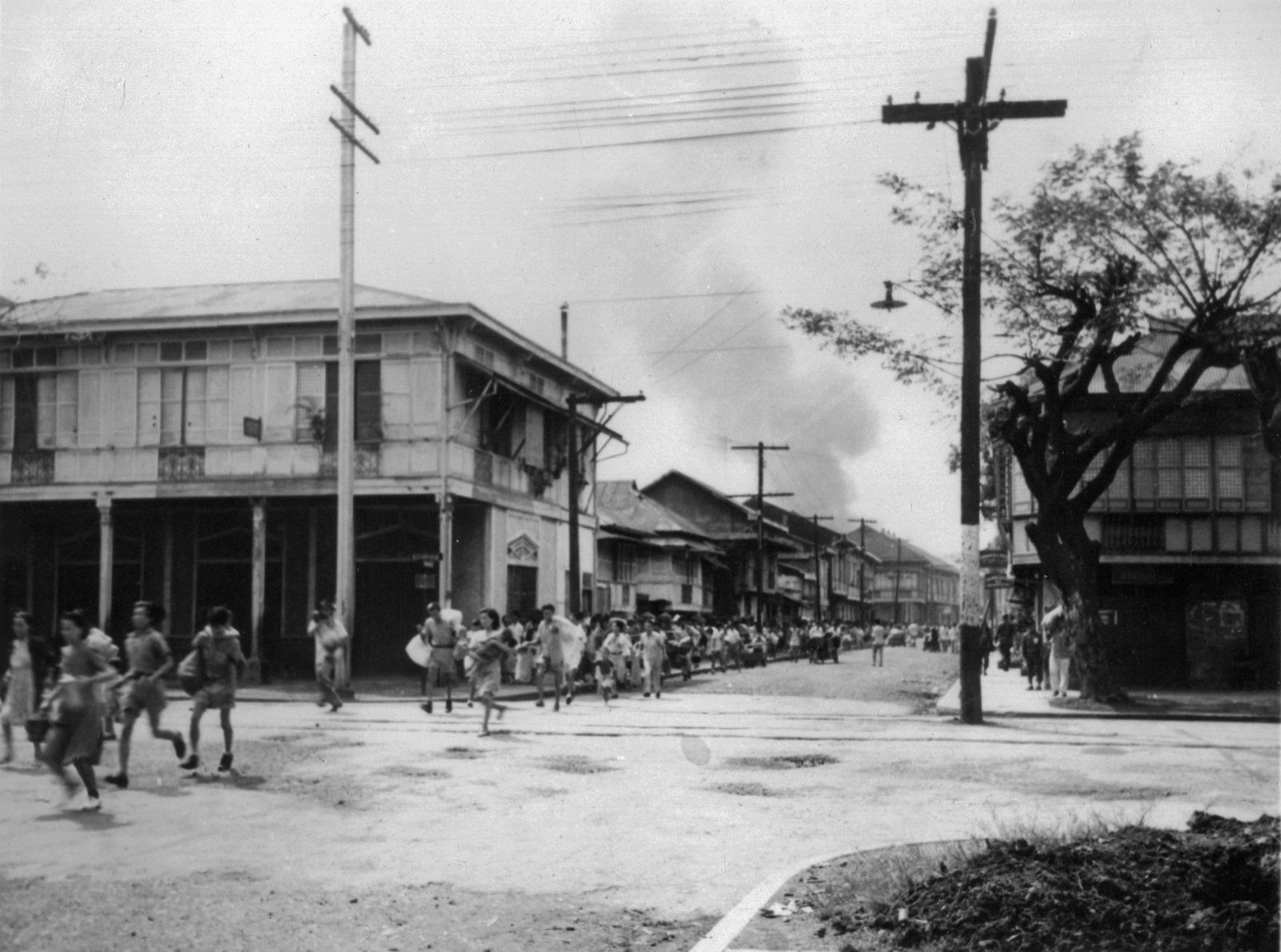 Black and white photograph: women and children run across a wide street lined with two-story buildings and telegraph poles, while a column of people hurries along the road behind them and a tall cloud of smoke rises over the rooftops.