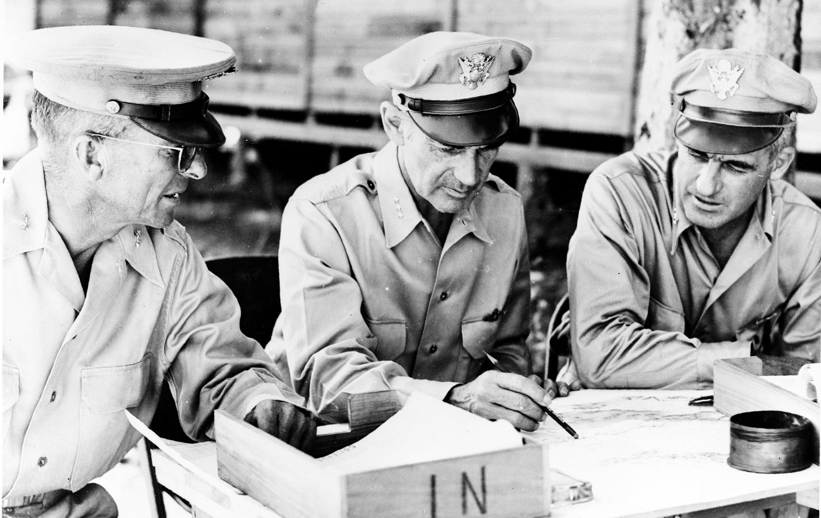 Black and white photograph of three American generals in khaki shirts and peaked caps seated at a field table, studying a map; the one in the middle holds a pencil over it.