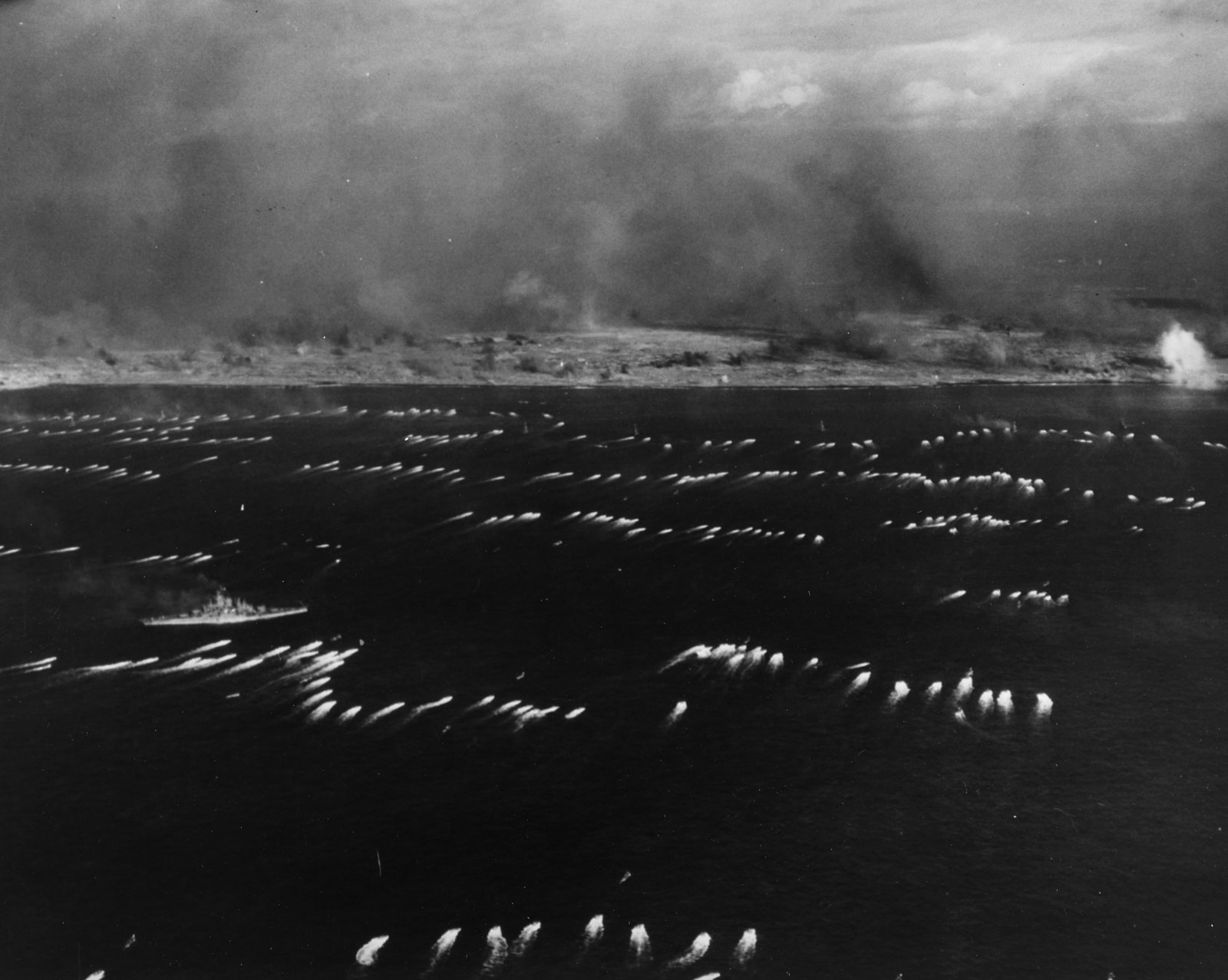 Black and white aerial photograph: rows of amphibious tractors trail white wakes across a dark sea toward a low island shore hidden under a huge cloud of smoke and dust, with a battleship on the left and a shell burst on the right.