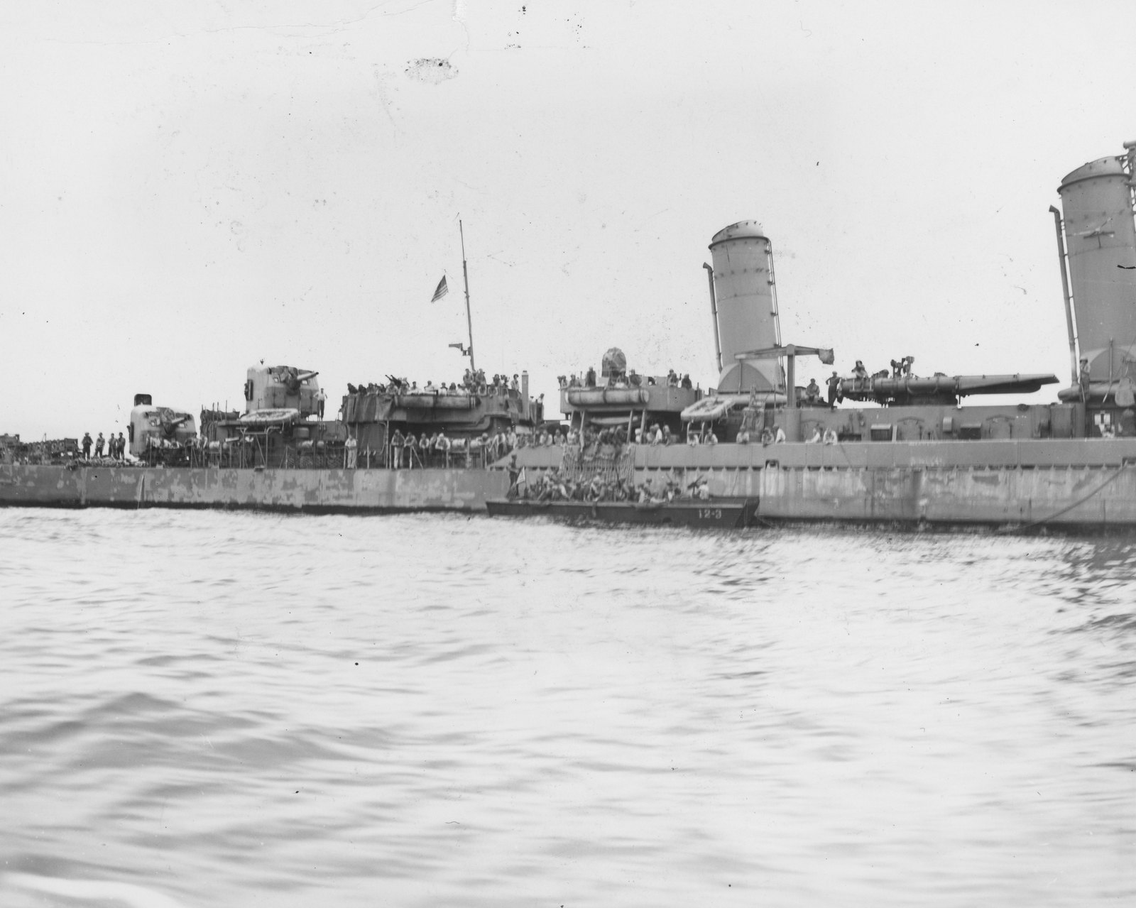 Black-and-white photograph of American cavalrymen with rifles and packs climbing down from the side of a destroyer into a landing craft crowded with soldiers.