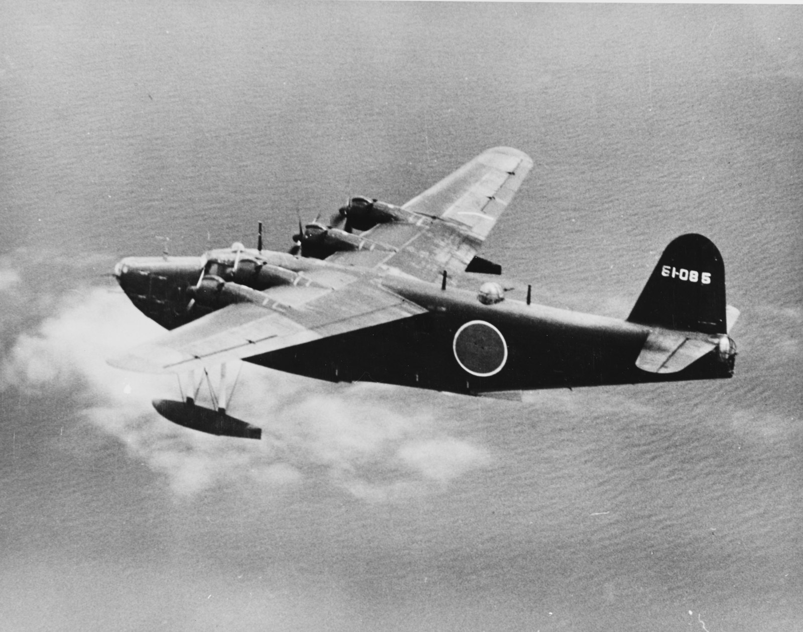 Black and white photograph of a large four-engined Japanese flying boat with a dark hull and a red sun roundel on its side, flying low over the sea with clouds below.