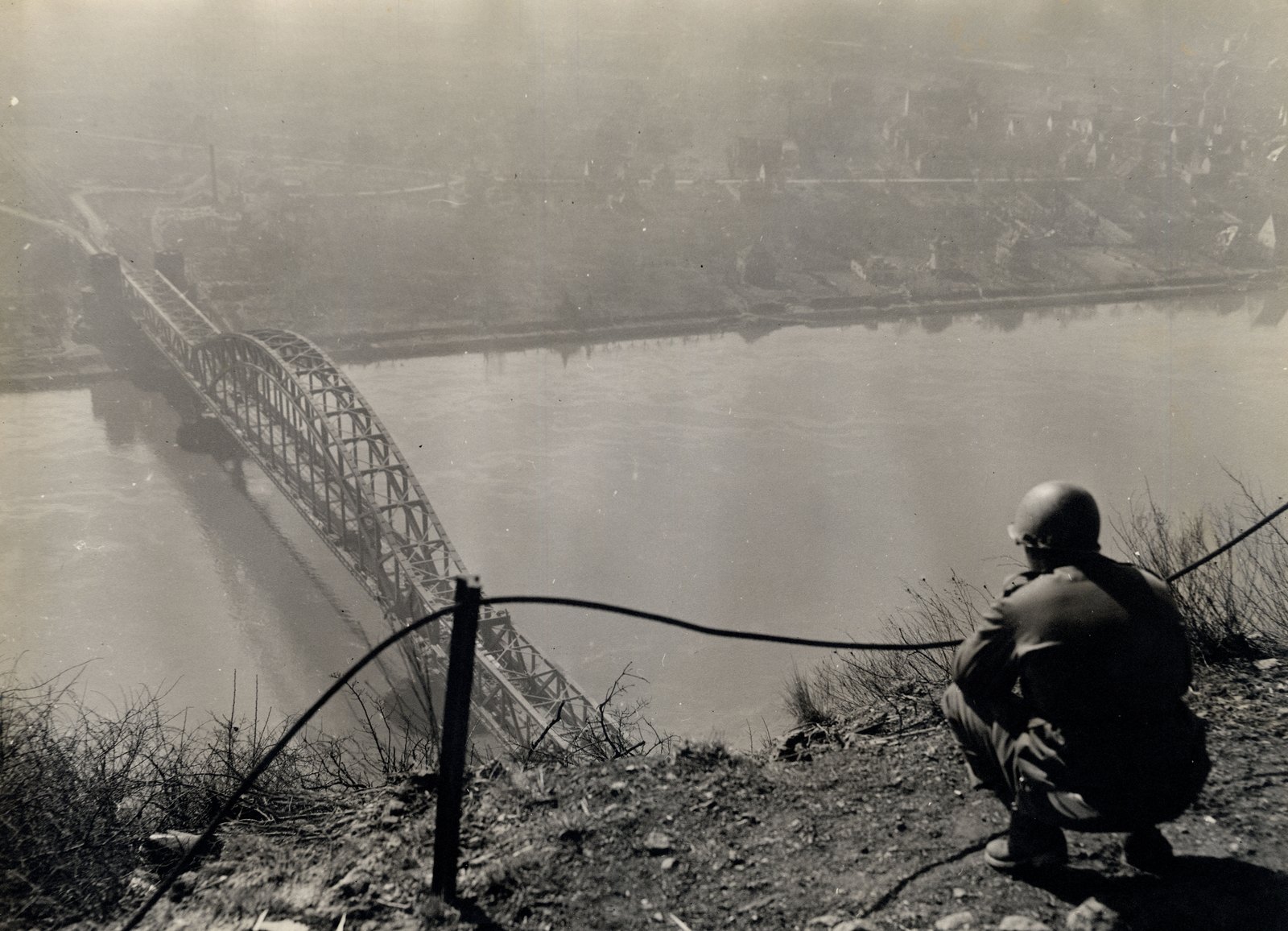 Black and white photograph: an American soldier in a helmet crouches on a rocky hilltop and looks down at a long steel arch railway bridge crossing a wide river, with a town on the far bank in haze.