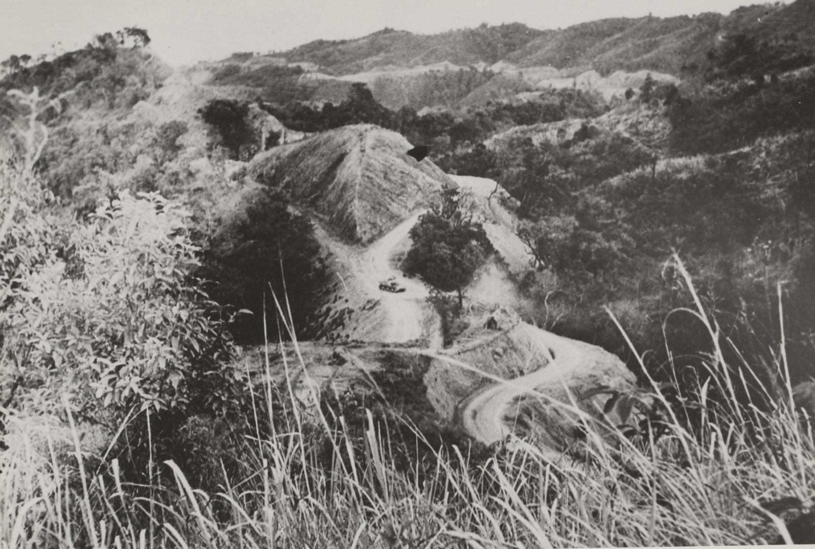 Black and white photograph: a narrow dirt road winds in tight hairpin bends through steep, scrub-covered hills, with a single tank on one of the bends and tall grass in the foreground.