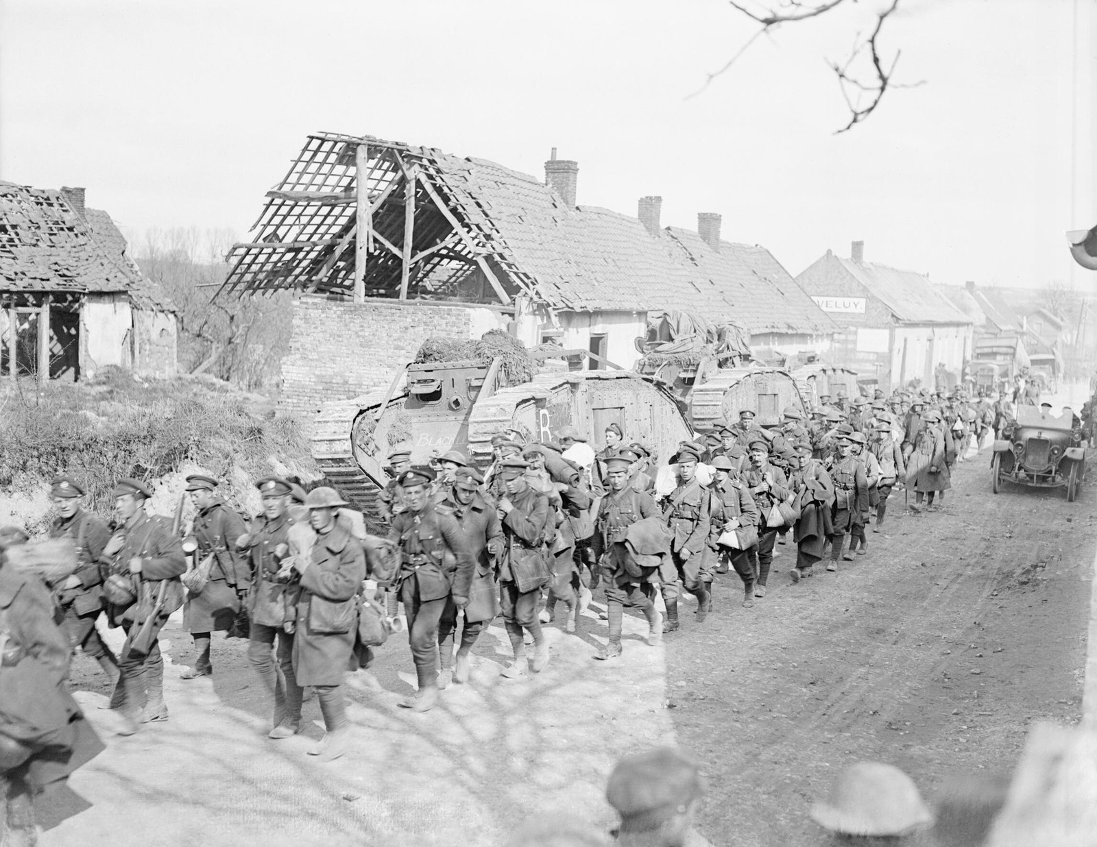 Black-and-white photograph of a long column of British infantry with packs and greatcoats marching down a village street past a row of camouflaged Mark V tanks and houses with smashed roofs.