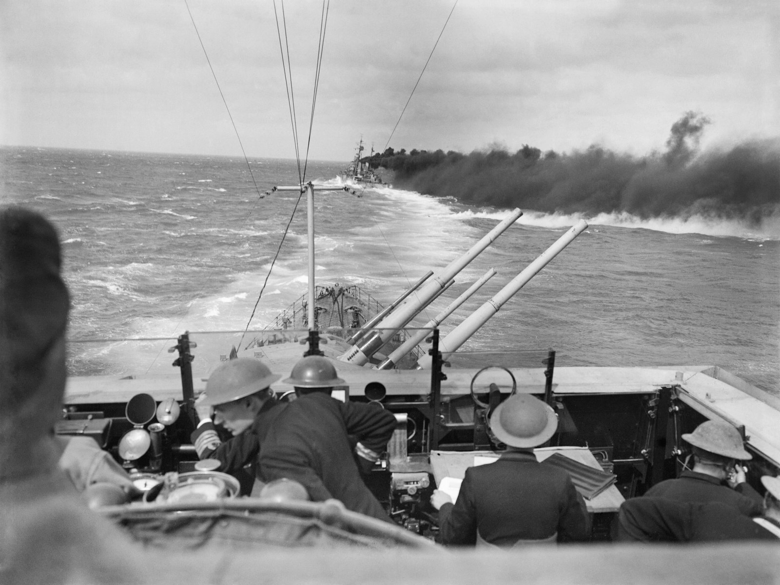 Black-and-white photograph from the bridge of a British cruiser in a rough sea: officers in steel helmets in the foreground, raised twin gun barrels on the forecastle, and ahead a second cruiser trailing a long bank of black smoke across the water.
