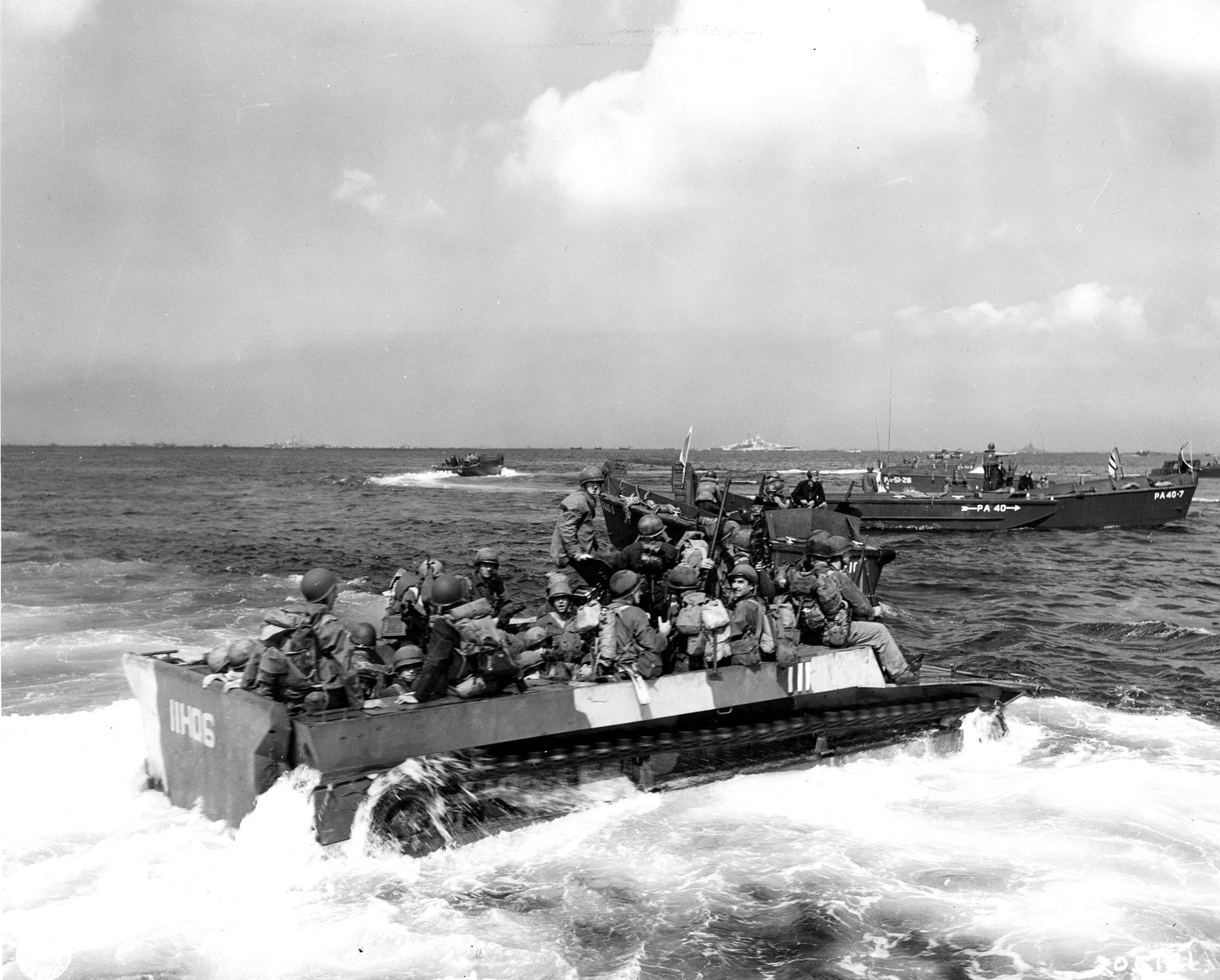 Black and white photograph: soldiers in helmets packed into an open amphibian tractor numbered 11-06 churn through choppy water toward the shore, with landing craft and distant warships behind them.
