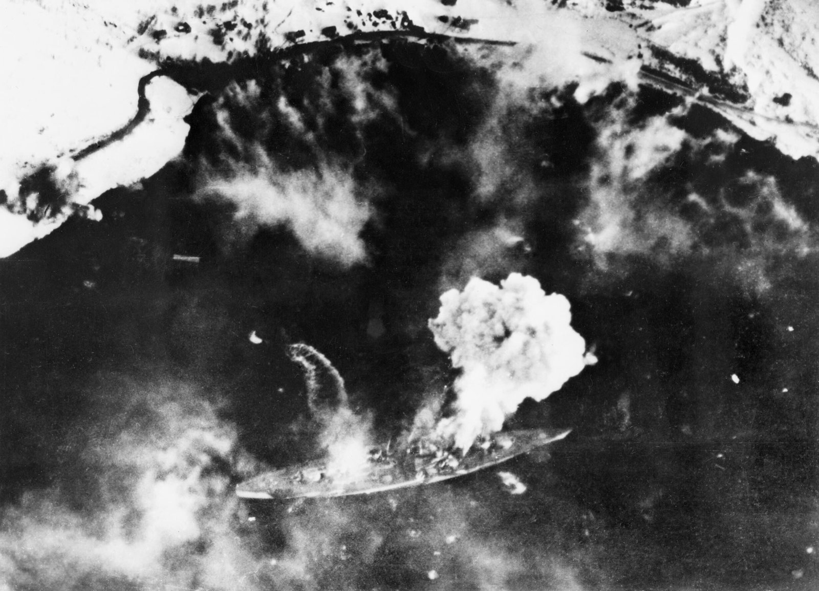 Black and white aerial photograph looking down into a fjord with snowy shores: a long battleship lies near the bottom of the frame while a large bomb explosion billows smoke from her middle and drifting smoke covers the dark water.