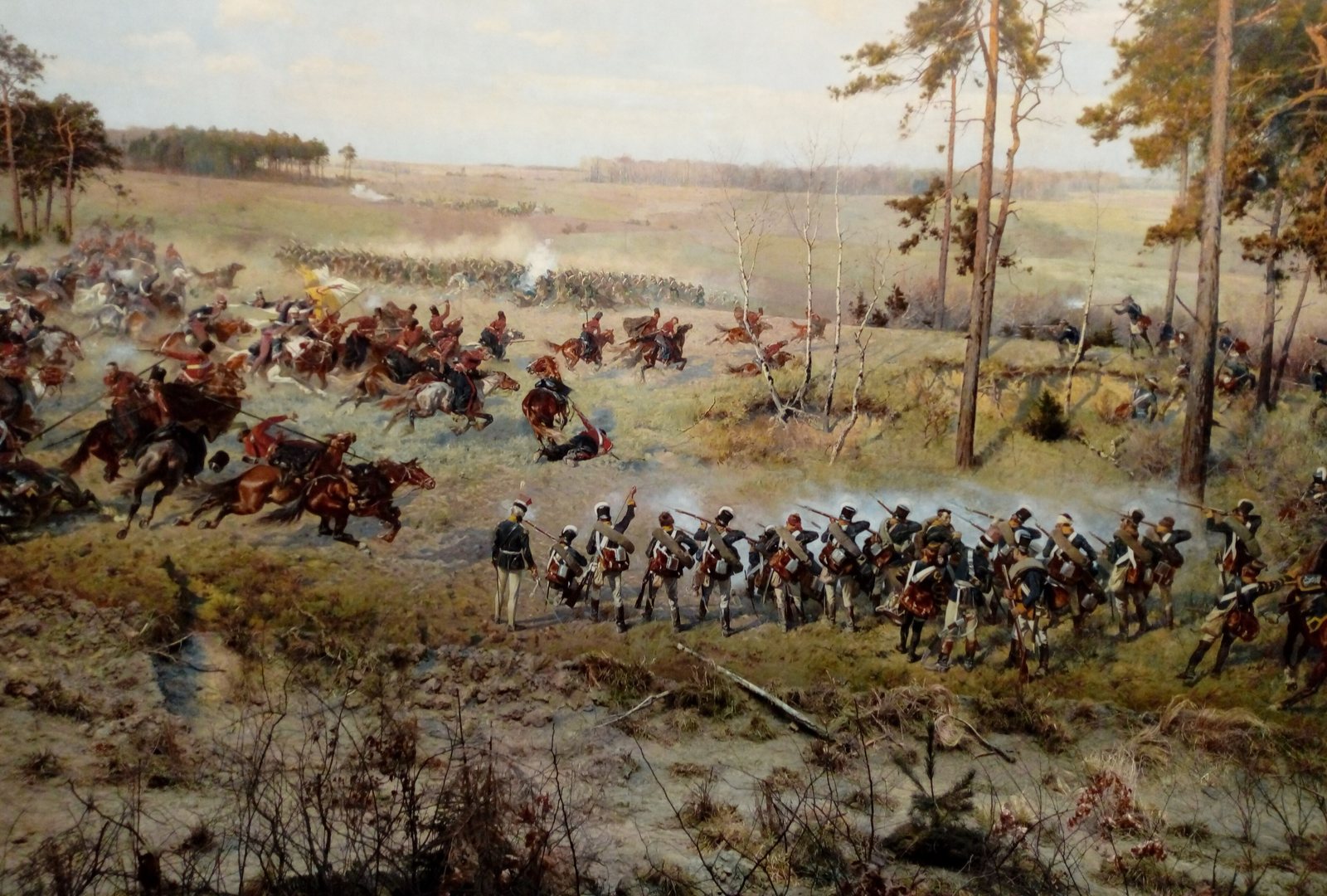 Detail of a huge painted panorama: a line of infantry in dark coats and white trousers advances across rough heath beside tall pines, while cavalry in red and blue gallops across the open field behind them under a pale sky.