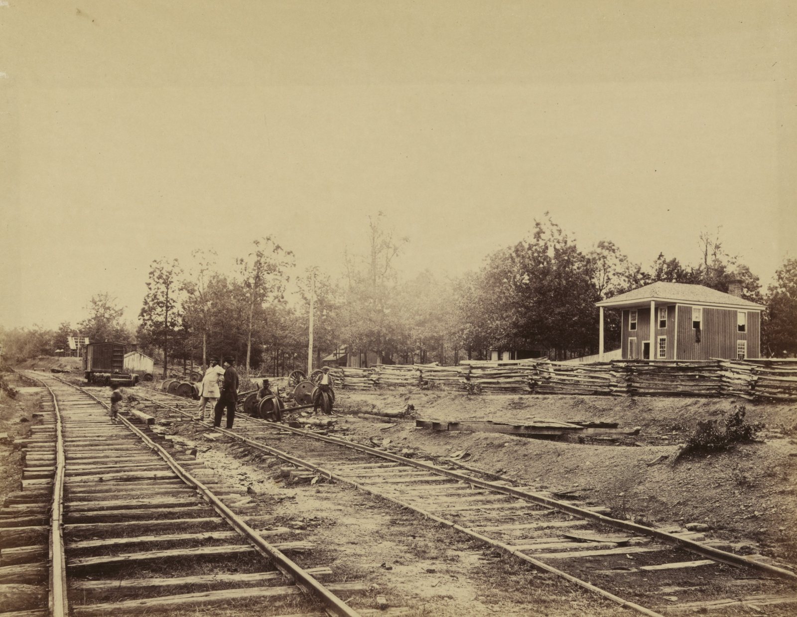 Sepia photograph of a rural railroad station: two sets of tracks run toward a small two story wooden depot with a porch, behind a long split rail fence, while a few men stand by railroad wheels on the line and trees rise behind.