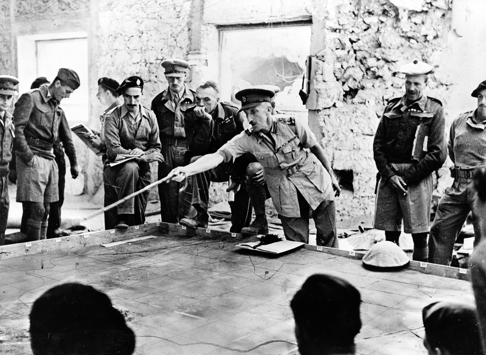 Black and white photograph: in a ruined stone building, a British officer in a peaked cap points with a long stick at a sand table model on the floor while a ring of officers in berets, caps and shorts watches him.