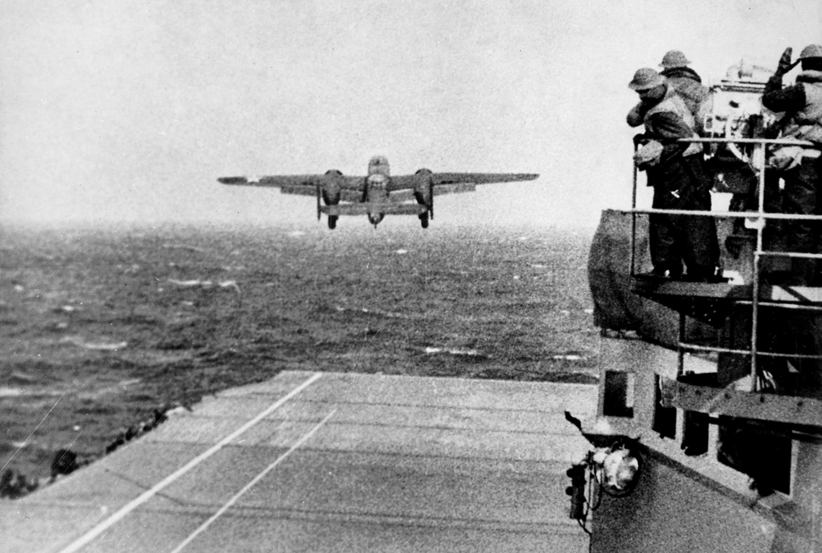 Black and white photograph: a twin-engine B-25 bomber lifts off over the bow of the carrier USS Hornet above a rough gray sea, while sailors in helmets and foul-weather gear watch from a gun platform on the right.
