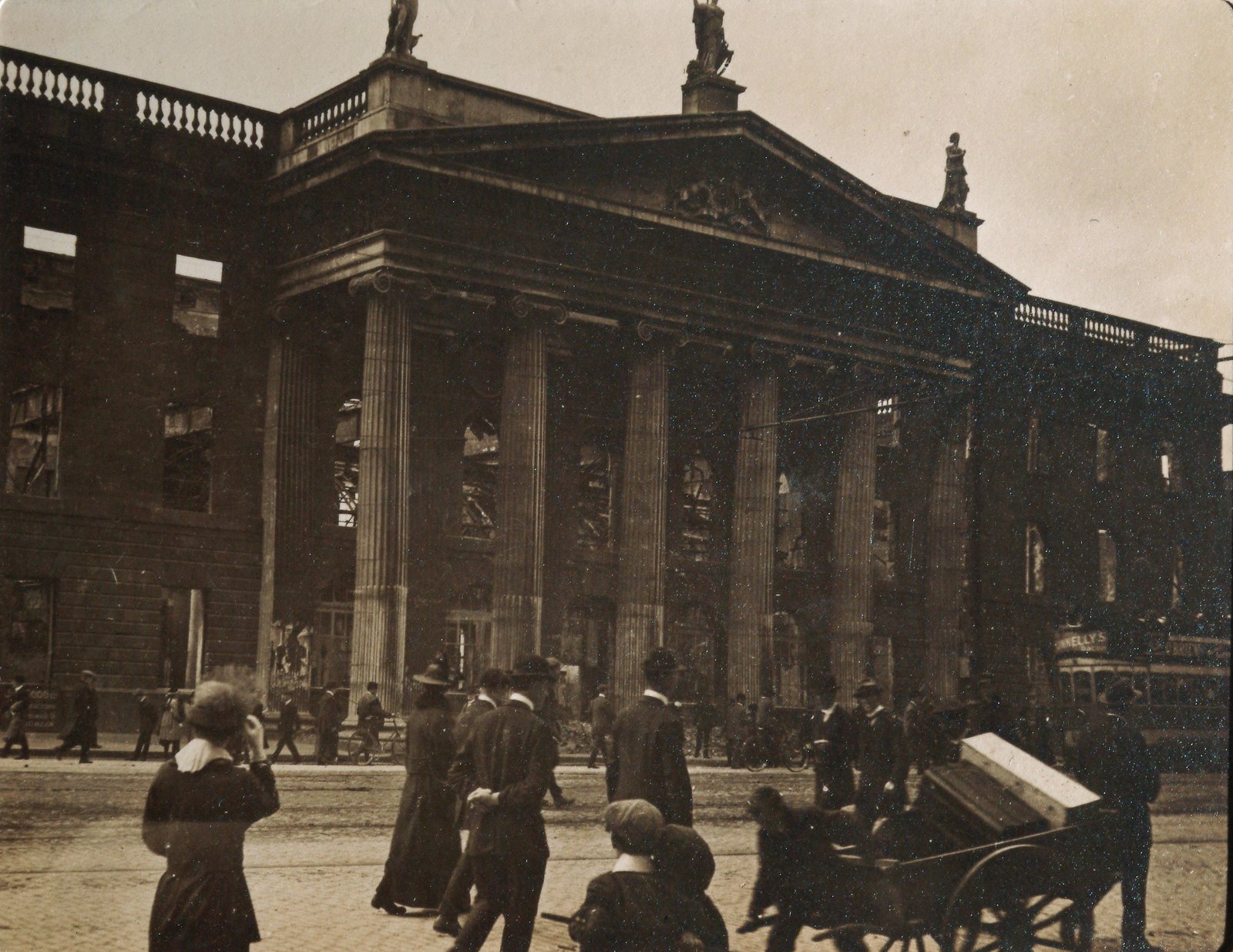 Sepia photograph of the General Post Office in Dublin in May 1916: the classical portico still stands, but the windows behind the columns are empty and the building is a burned-out shell, while people in hats and long coats walk past on the street in front.