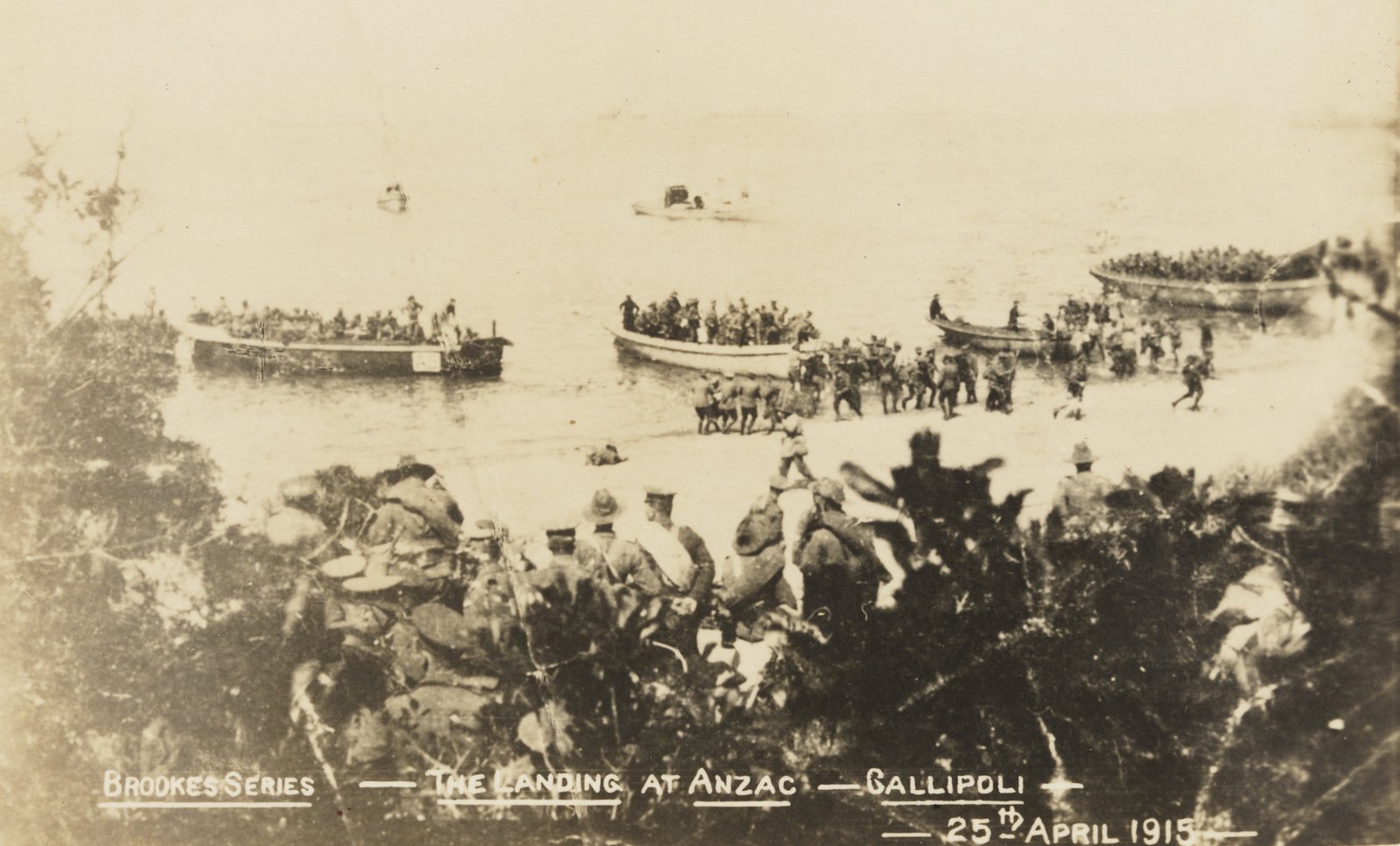 Faded sepia photograph taken from scrub on a hillside: rowing boats crowded with soldiers lie off a narrow beach while men wade ashore and run up the sand, with a caption along the bottom reading The Landing at Anzac, Gallipoli, 25th April 1915.