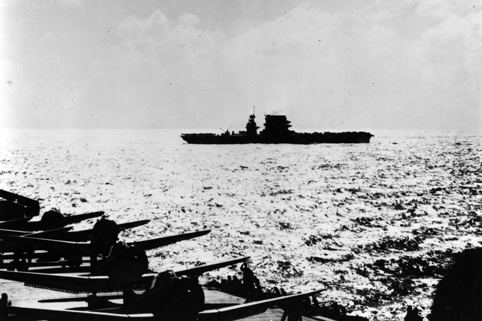 Black and white photograph taken from a carrier deck: the dark silhouette of the aircraft carrier USS Lexington sails across a glittering sea under a pale sky, while the folded wings of dive bombers fill the lower left corner.