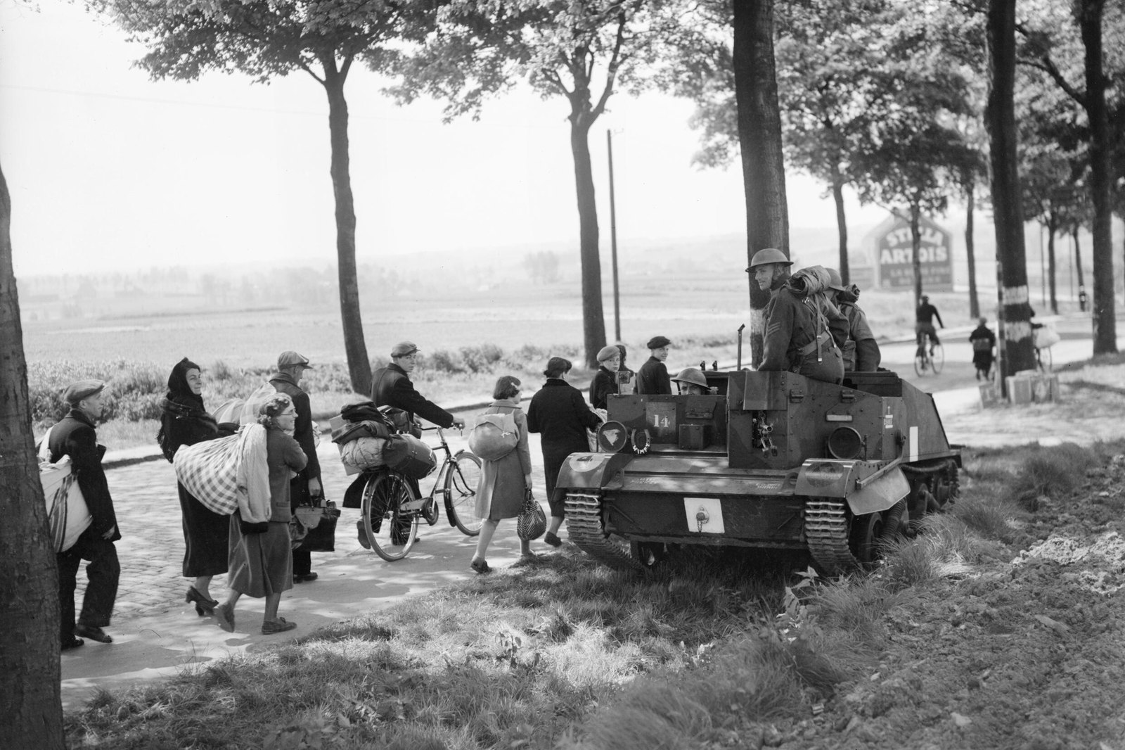 Black and white photograph of a tree-lined country road: a small British tracked Bren gun carrier with helmeted soldiers drives past Belgian refugees on foot and on bicycles who carry bundles and bags, with flat fields behind them.