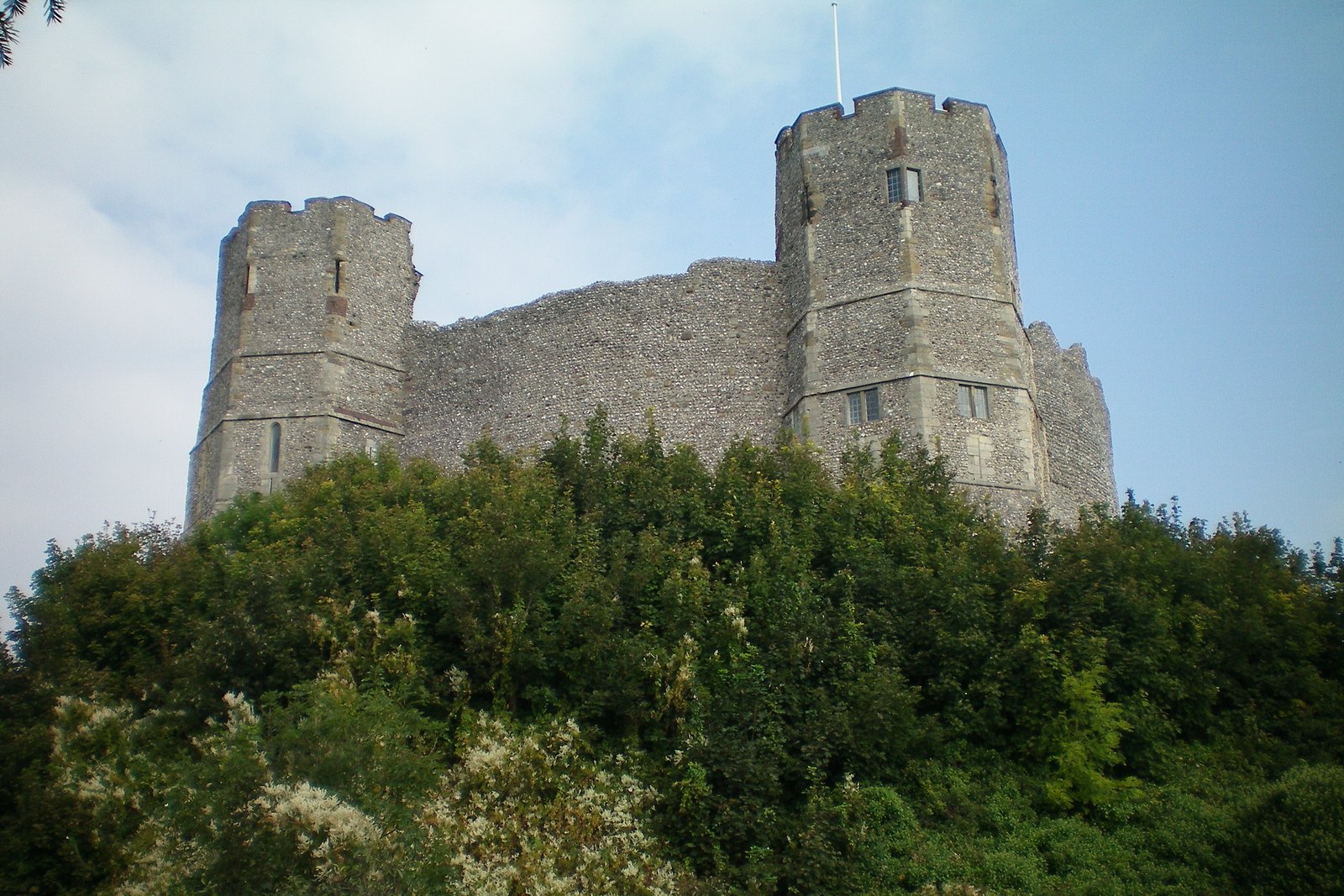 Color photograph of a medieval castle keep on a mound: a high wall of gray flint joins two tall many-sided towers with battlements and small windows, rising above a thick bank of green trees and bushes against a pale blue sky.