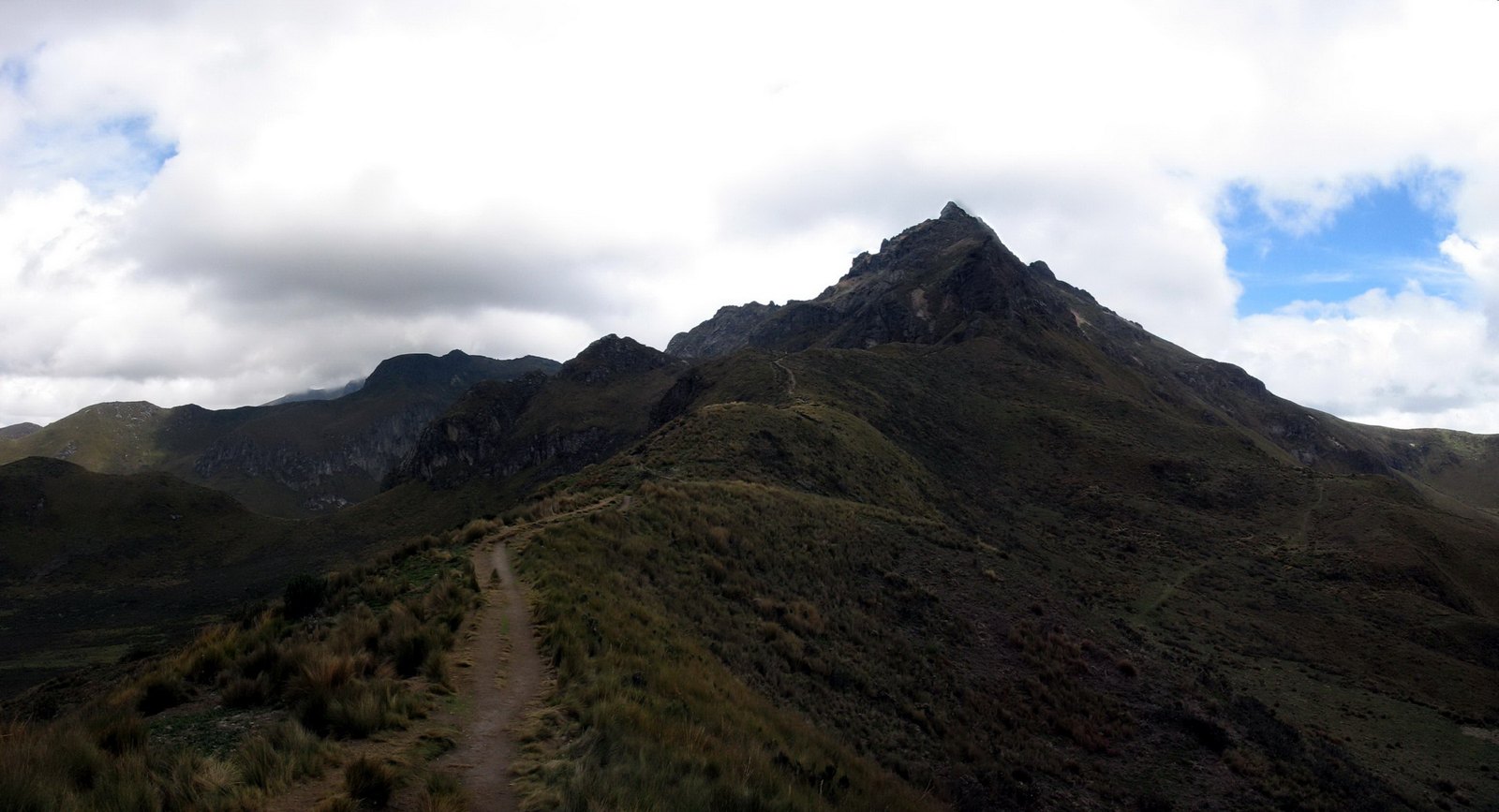 A grassy mountain ridge with a narrow footpath climbing toward the dark rocky peak of Rucu Pichincha under heavy white clouds and patches of blue sky.