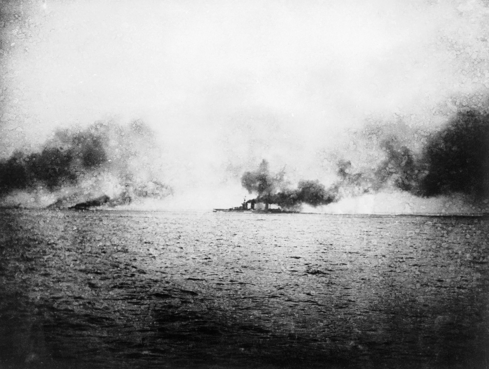 Grainy black and white photograph taken at sea during the battle: a gray sea in the foreground, and on the horizon a battlecruiser almost hidden by dark smoke and the splashes of shells, with more smoke drifting across the line.