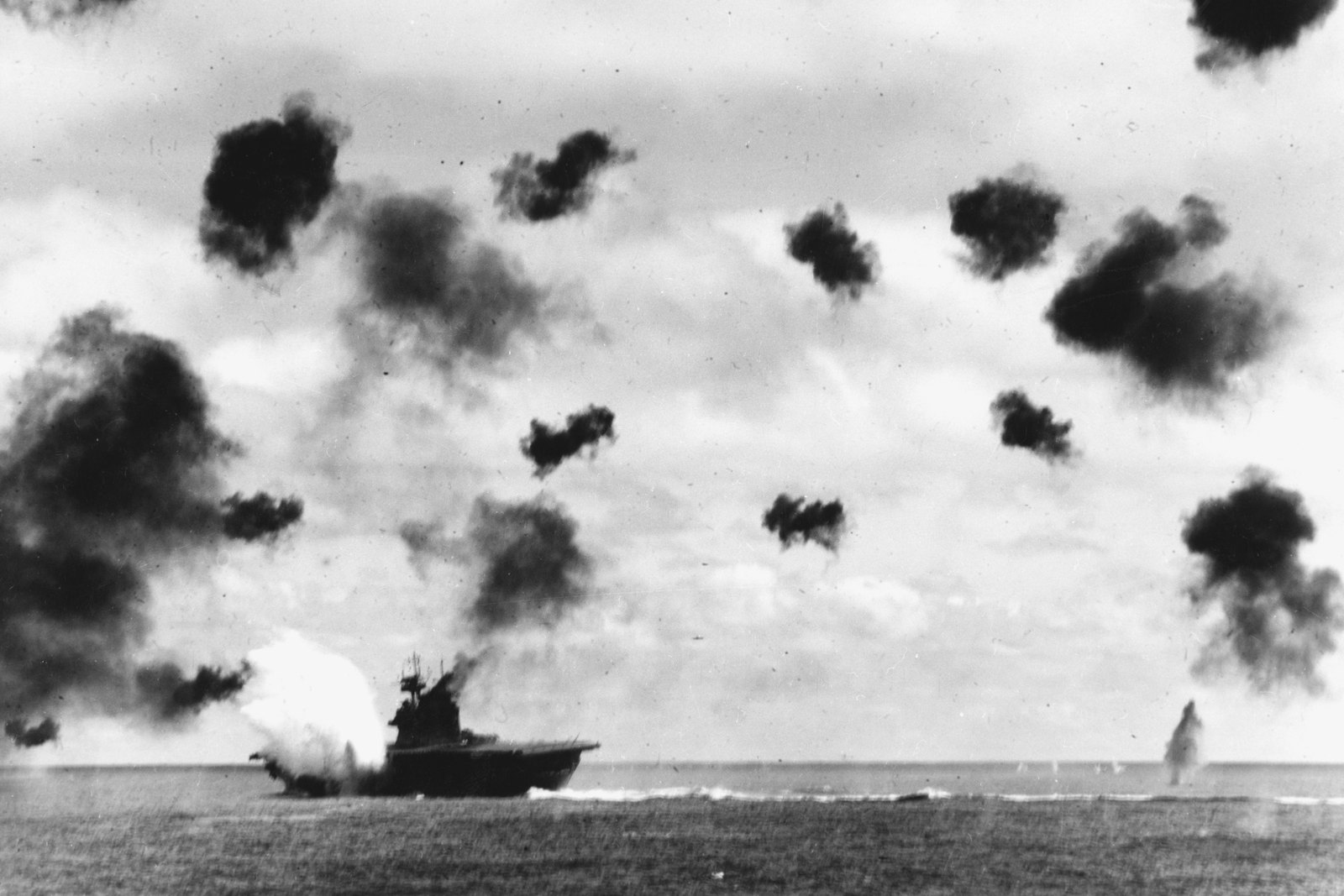 Black and white photograph of the aircraft carrier USS Yorktown on the horizon at the moment a torpedo strikes her side, throwing up a white burst of water, while dark puffs of anti-aircraft fire fill the sky above.