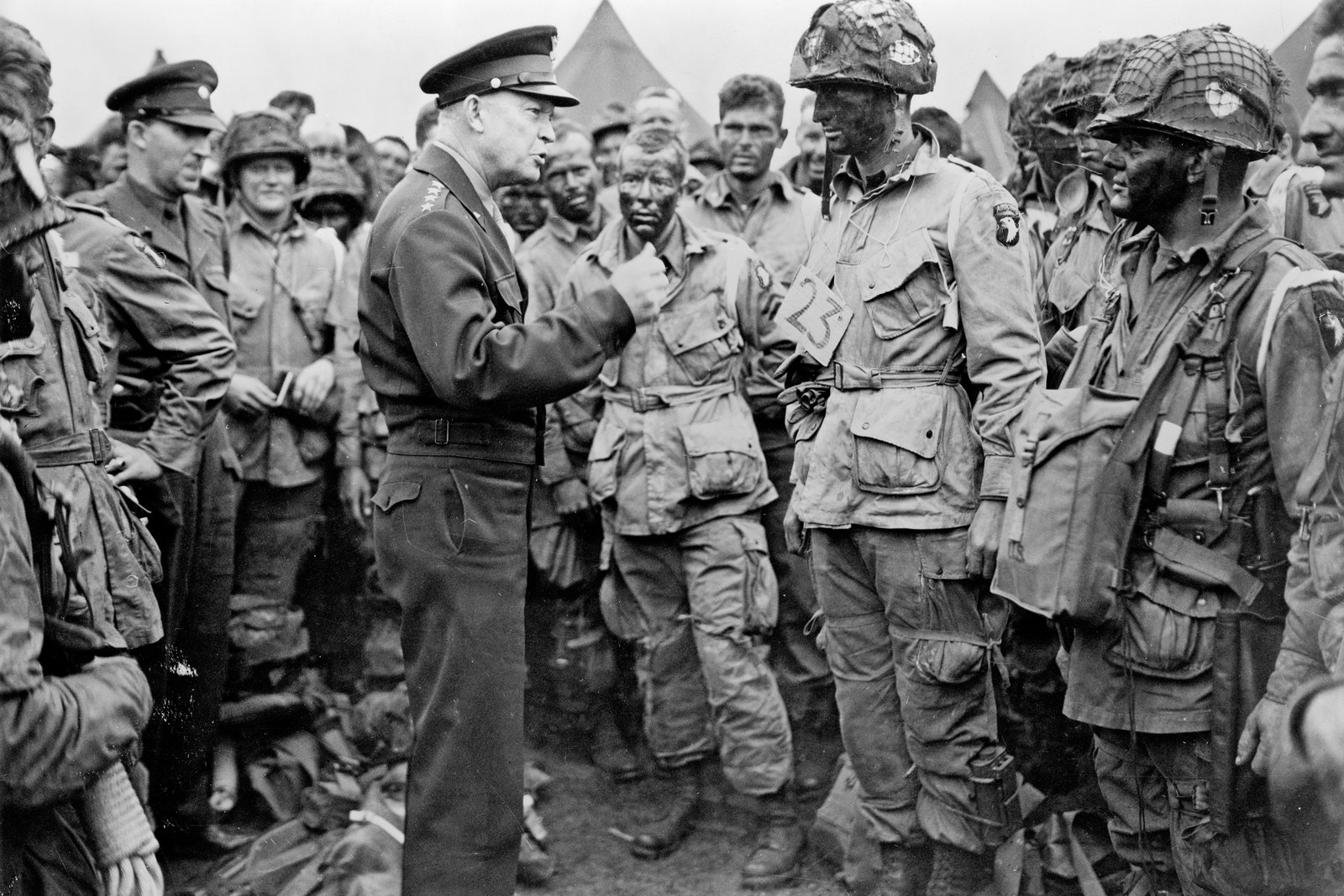 Black and white photograph of General Eisenhower in uniform and peaked cap talking with a crowd of American paratroopers in jump uniforms and netted helmets, their faces blackened, one with a number 23 card on his chest, with tents in the background.