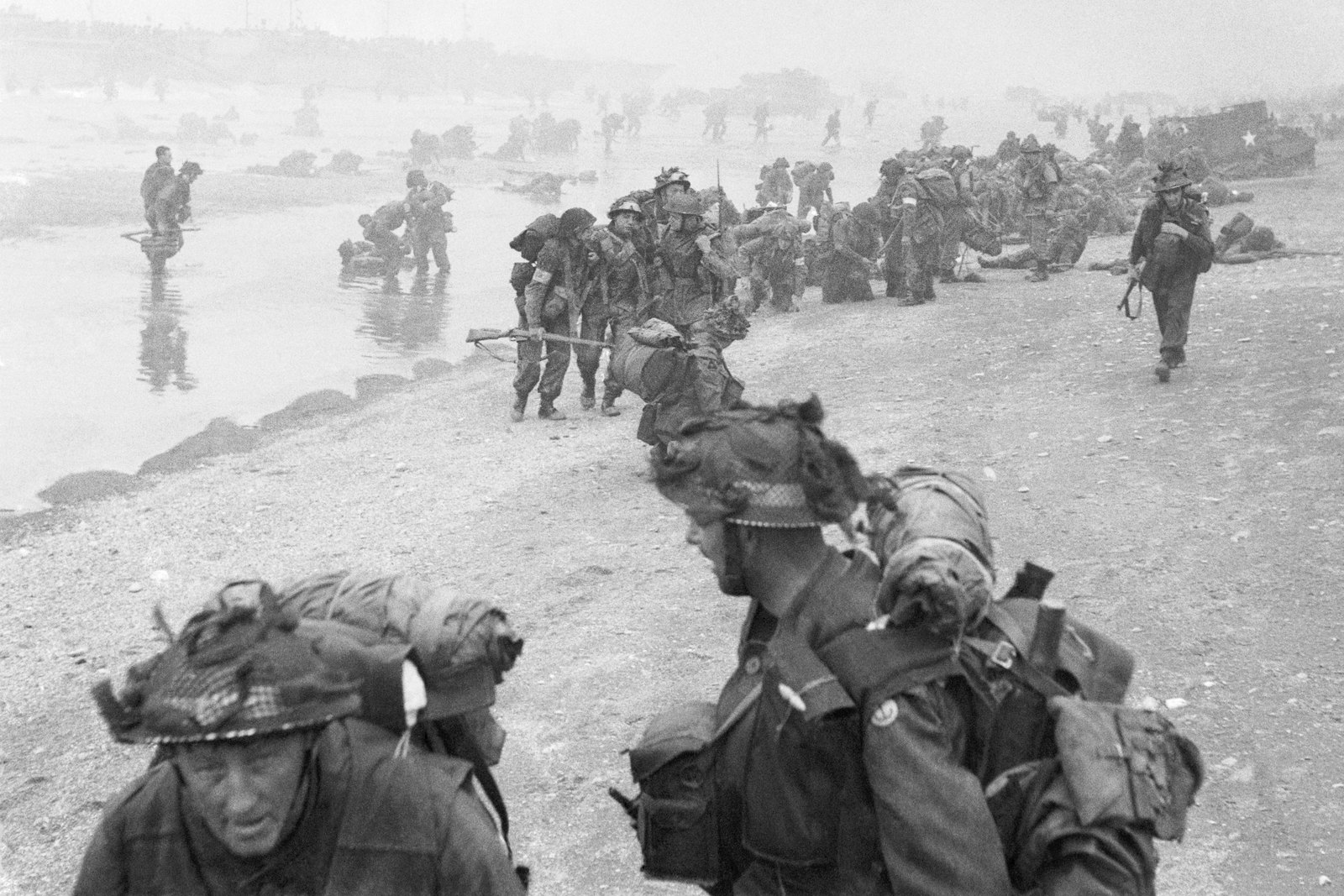 Black and white photograph of British soldiers with heavy packs and netted helmets on a misty beach, sappers in the foreground and medical orderlies helping wounded men behind them, with landing craft and vehicles at the water's edge.