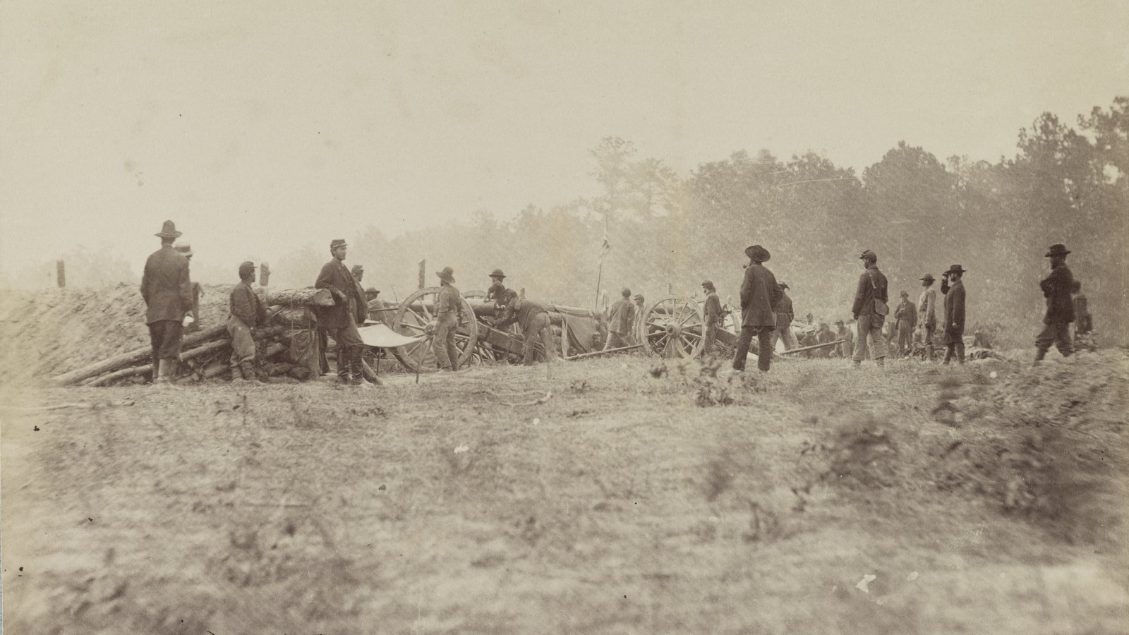 Faded sepia photograph of Union soldiers in slouch hats and kepis standing among captured cannon behind a low earthwork parapet of logs and soil, with smoky woods beyond and an open field in the foreground.