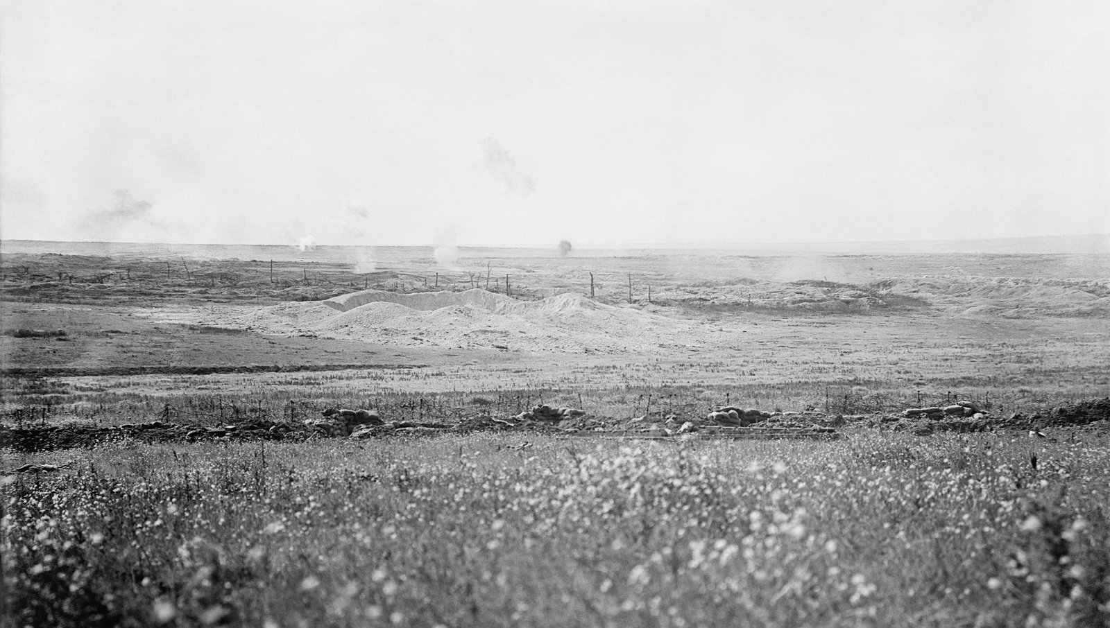 Black and white photograph: a flat, shell-torn landscape under a pale sky, with a long pale mound of chalk spoil from a mine crater in the middle distance, broken stakes of barbed wire, puffs of smoke on the horizon and a line of sandbags in the grass in the foreground.