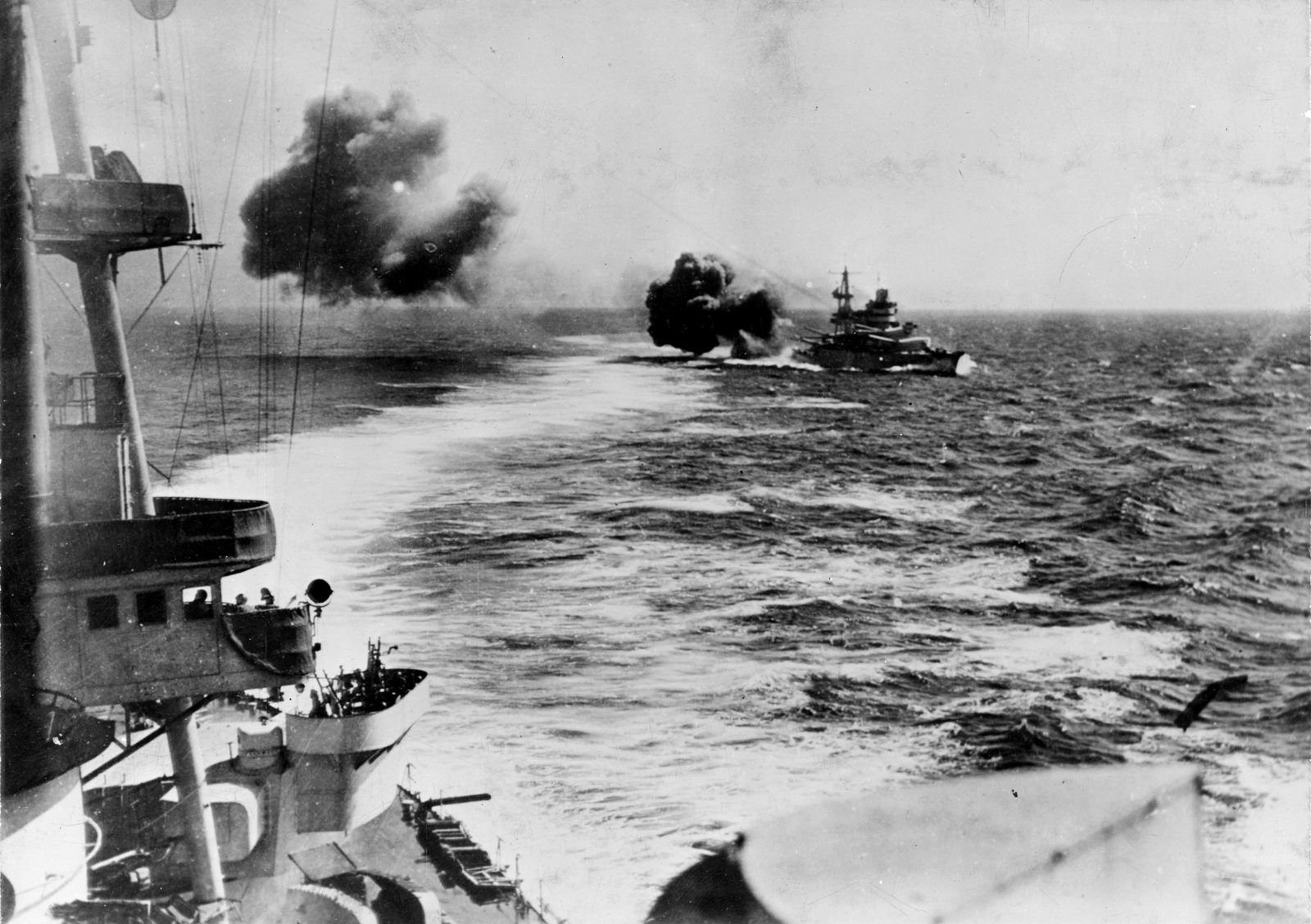 Black and white photograph taken from the deck of an Italian warship: ahead, across a choppy sea, the battleship Conte di Cavour fires a broadside, with two great clouds of dark gun smoke rolling away from her.