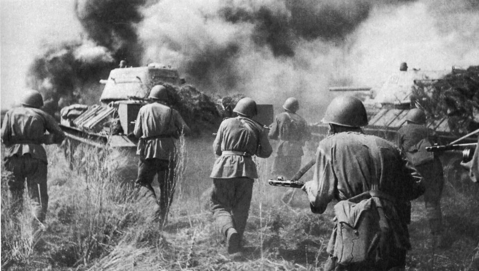 Black and white photograph: Soviet infantrymen in steel helmets, seen from behind, run forward through tall grass with submachine guns beside two T-34 tanks, under thick black and white smoke.