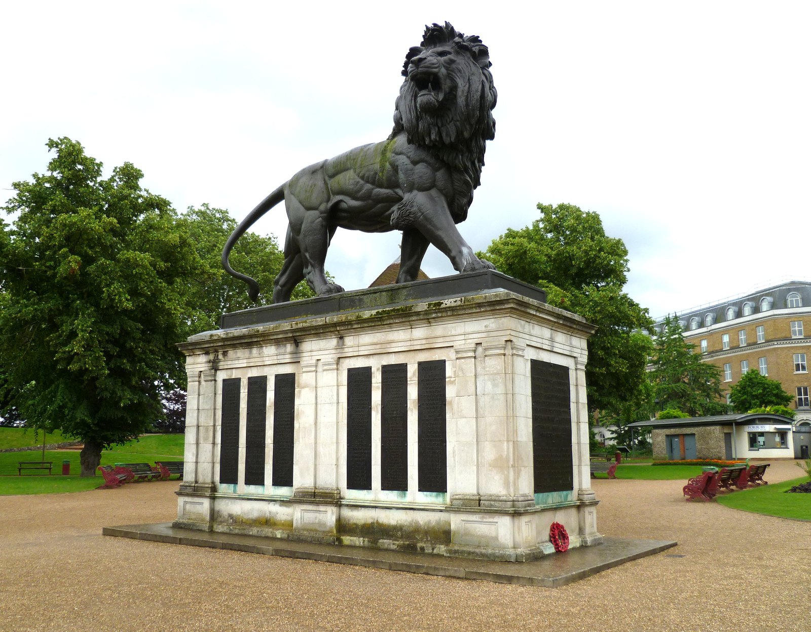 A huge cast-iron lion strides on top of a pale stone pedestal set with dark bronze panels listing names, a red poppy wreath at its base, in a public garden with trees, benches and a brick building behind.