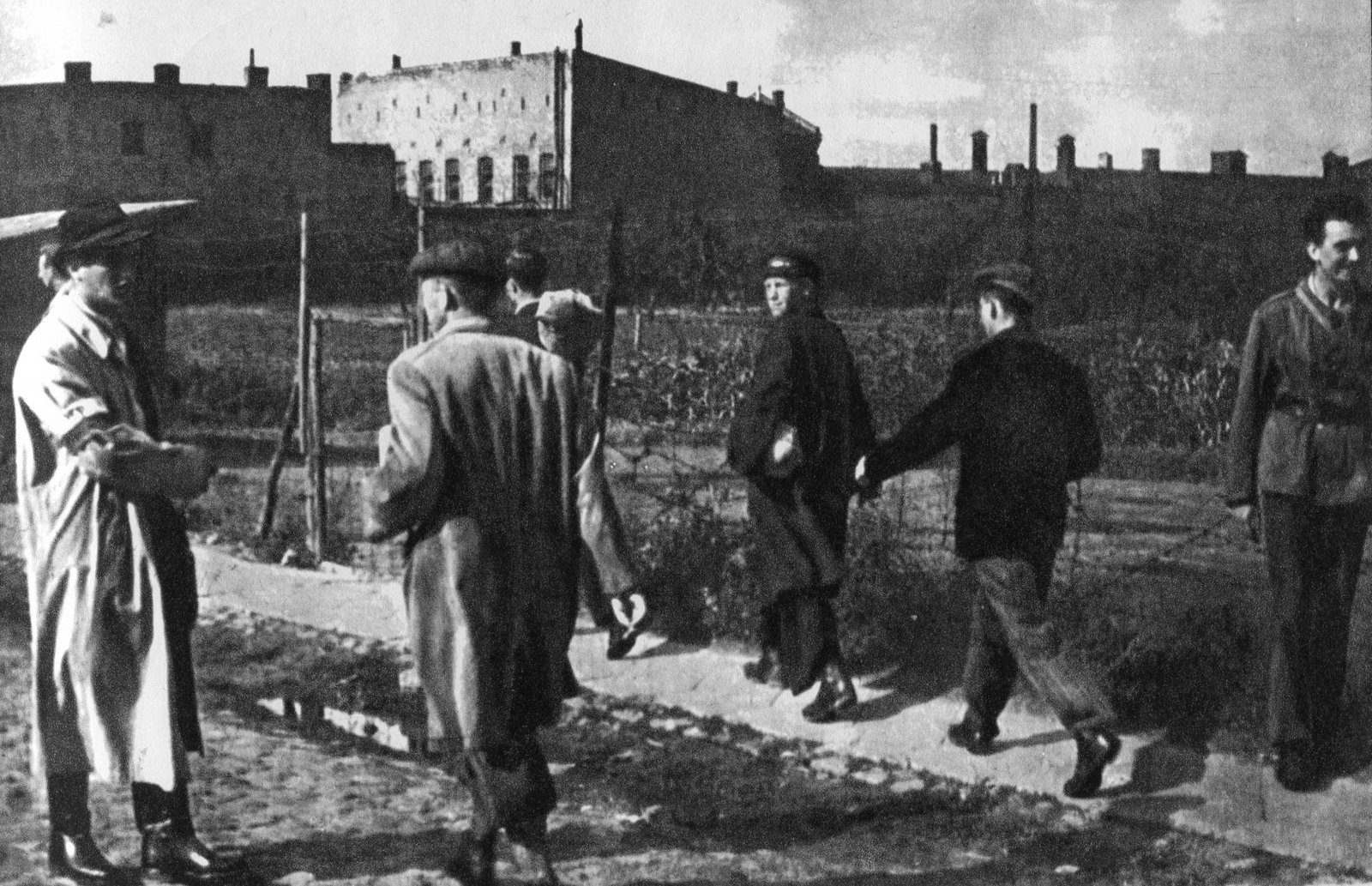 Black and white photograph of men in coats and caps walking along a cobbled street beside a wire fence in Wola, Warsaw, with brick factory buildings behind them; one man on the left in a light raincoat greets the others as they gather.