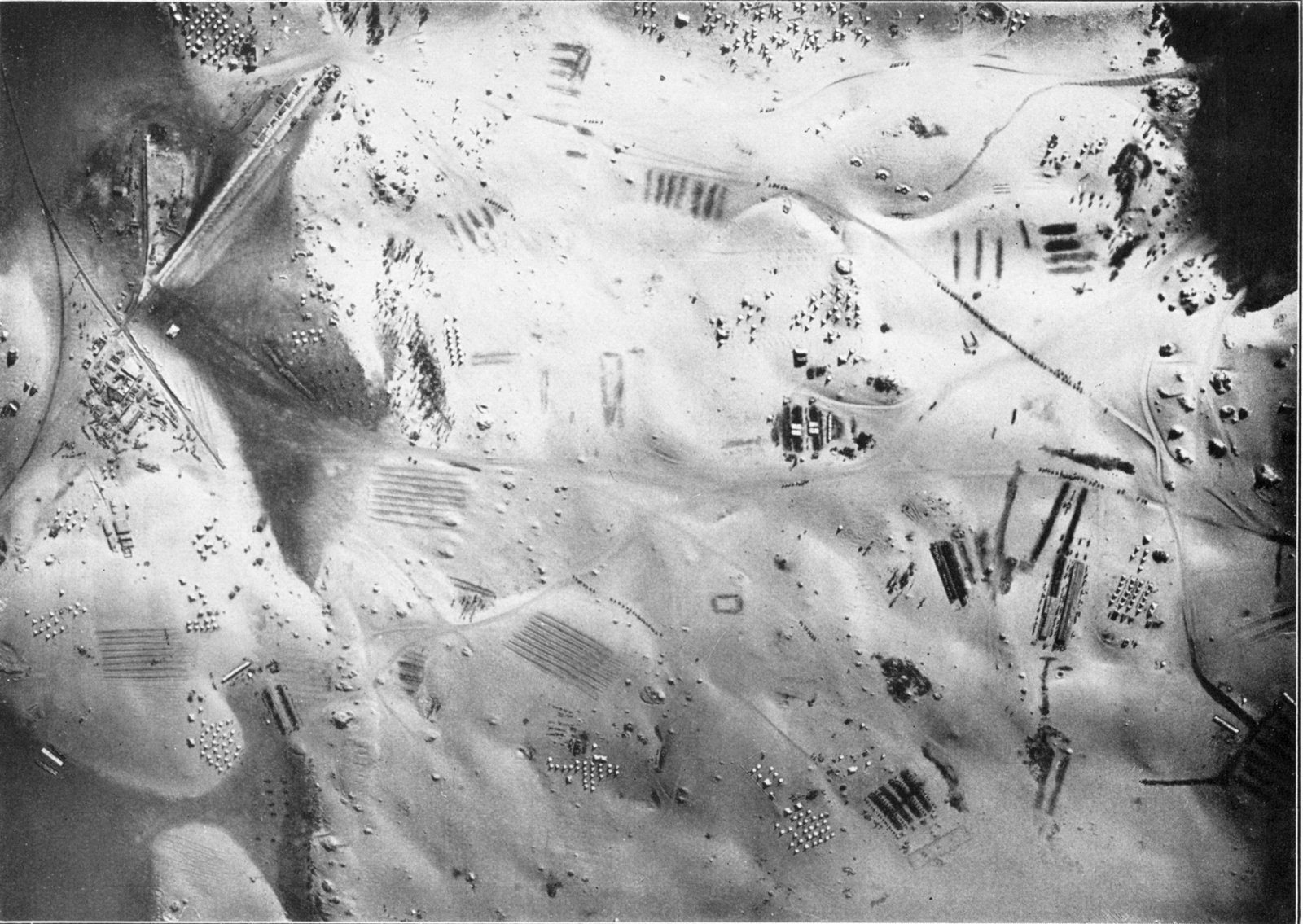 Black and white aerial photograph of a desert camp among pale sand dunes: rows of tents, horse lines, tracks and a railway line on the left, seen from directly above.