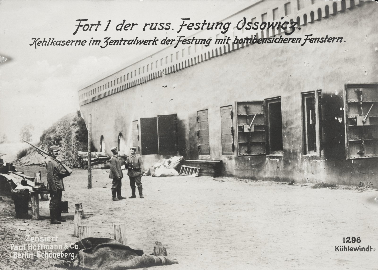 Black and white photograph of a long, low barracks wall of a fortress with heavy armored window shutters, three German soldiers in caps standing on the bare ground in front of it, and a German caption across the top reading Fort 1 der russ. Festung Ossowicz.