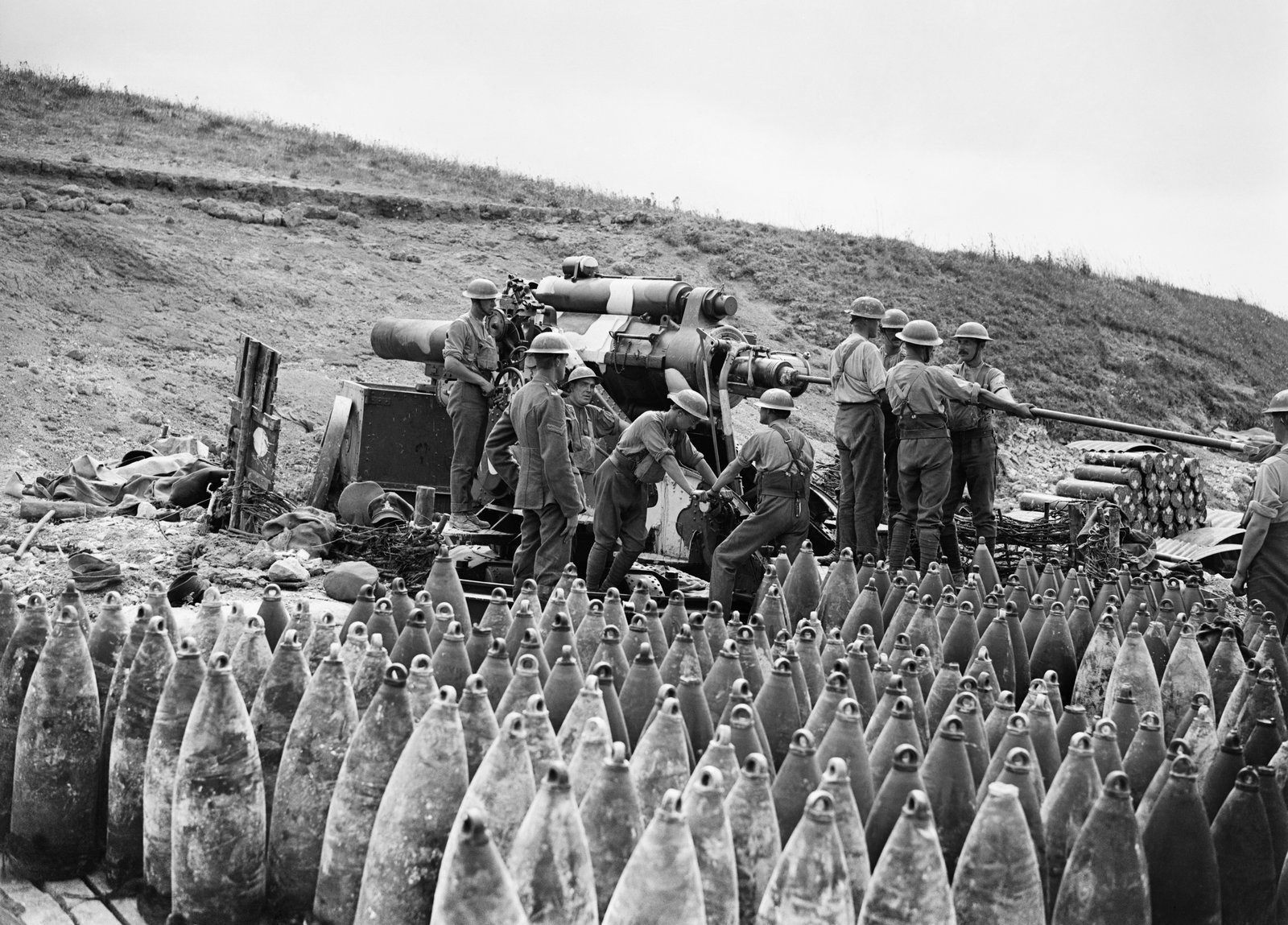Black and white photograph of British gunners in steel helmets and shirtsleeves loading a large howitzer on a chalky slope, with rows of hundreds of heavy shells standing upright in the foreground.