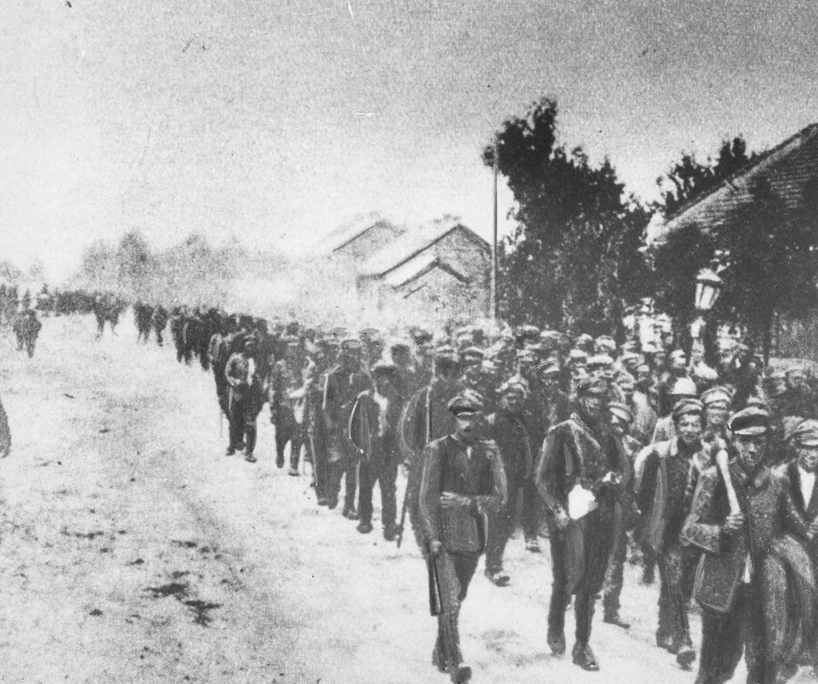 Grainy black and white photograph of a long column of men in caps and greatcoats marching down a dusty village street past low houses and trees, guarded by a soldier with a rifle in the foreground.