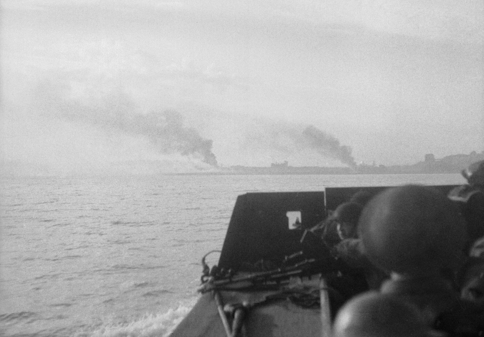 Black-and-white photograph taken from a landing craft heading for the Dieppe shore: the helmets of soldiers crouching in the boat fill the lower right corner, a machine gun rests on the bow, and across the gray water two columns of black smoke rise from the low coastline ahead.