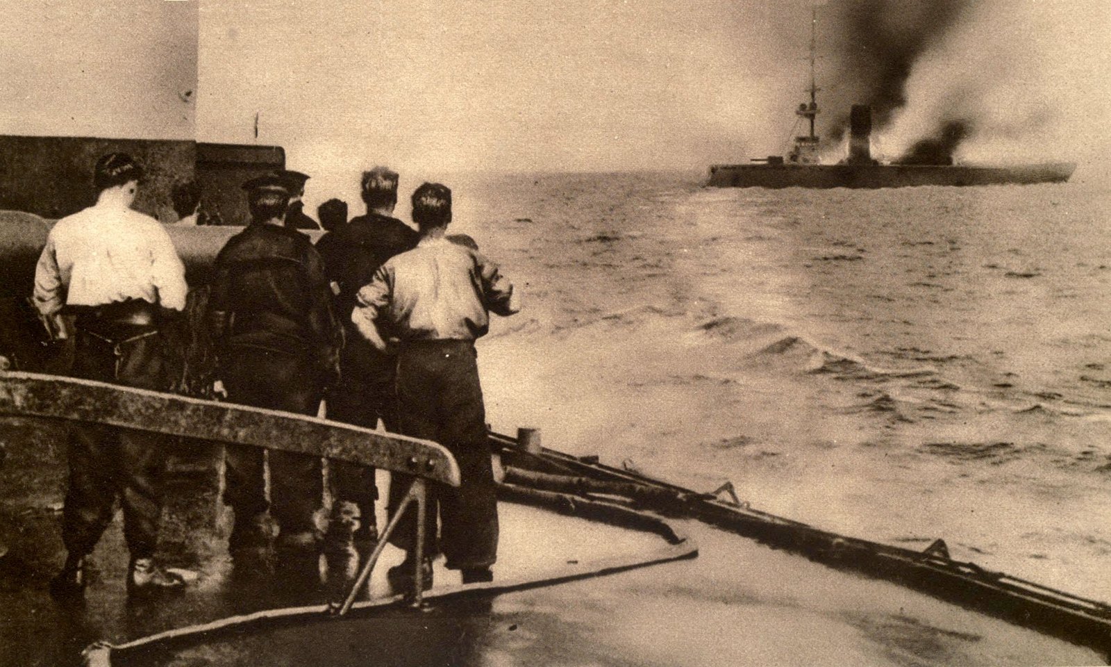 Sepia photograph taken from the deck of a British warship: sailors in shirtsleeves and caps stand by the rail watching a German cruiser in the distance, smoke pouring from her as she lies low in the water.
