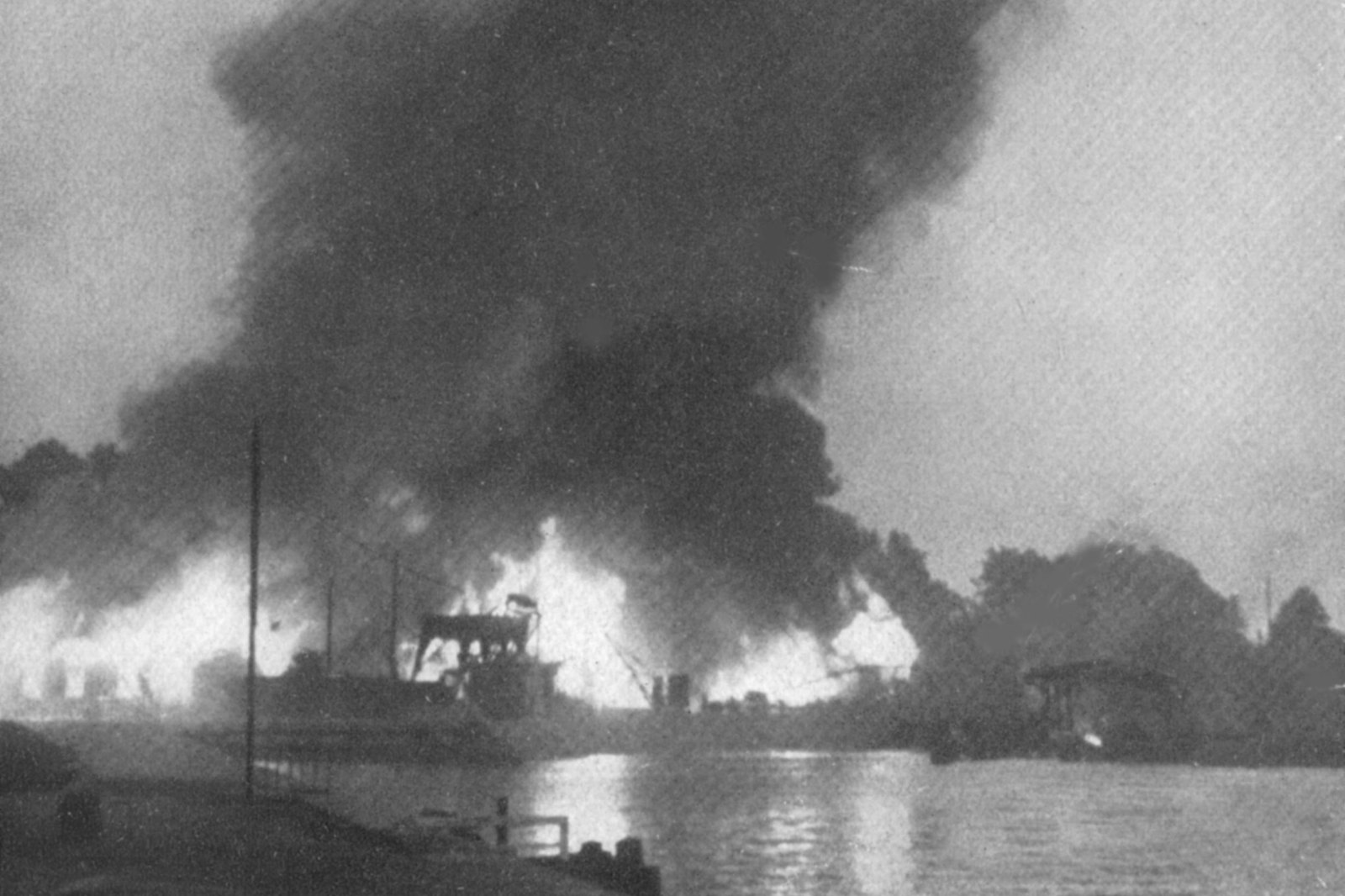 Black and white photograph: a huge column of black smoke and bright flames rise from harbor buildings on the far bank of a canal, with a crane and the hull of a ship in silhouette and still water in the foreground.