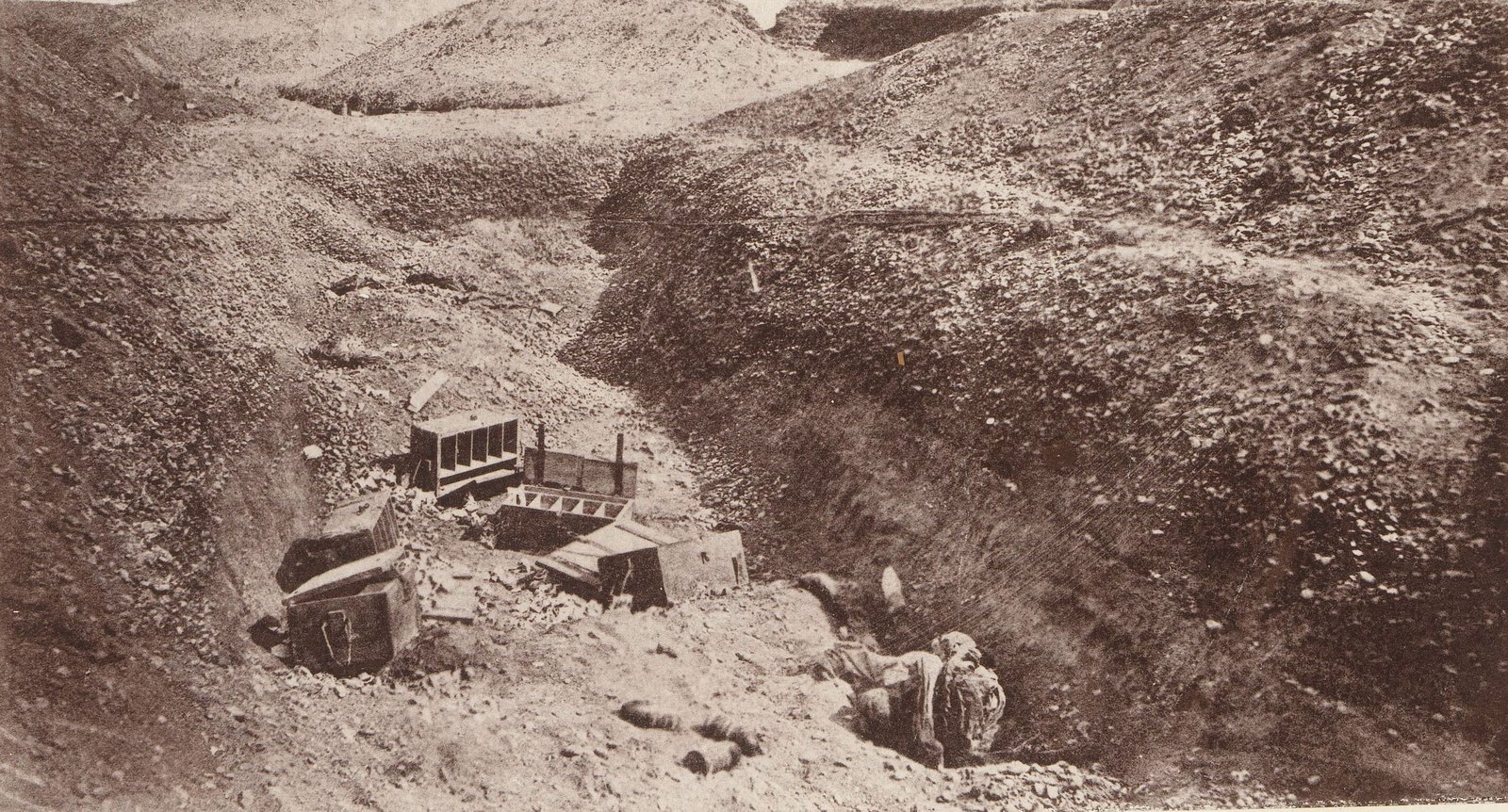 Sepia photograph of a deep, stony trench cut through desert earthworks, with broken ammunition boxes scattered on the floor and the body of a fallen soldier lying at the bottom of the bank.