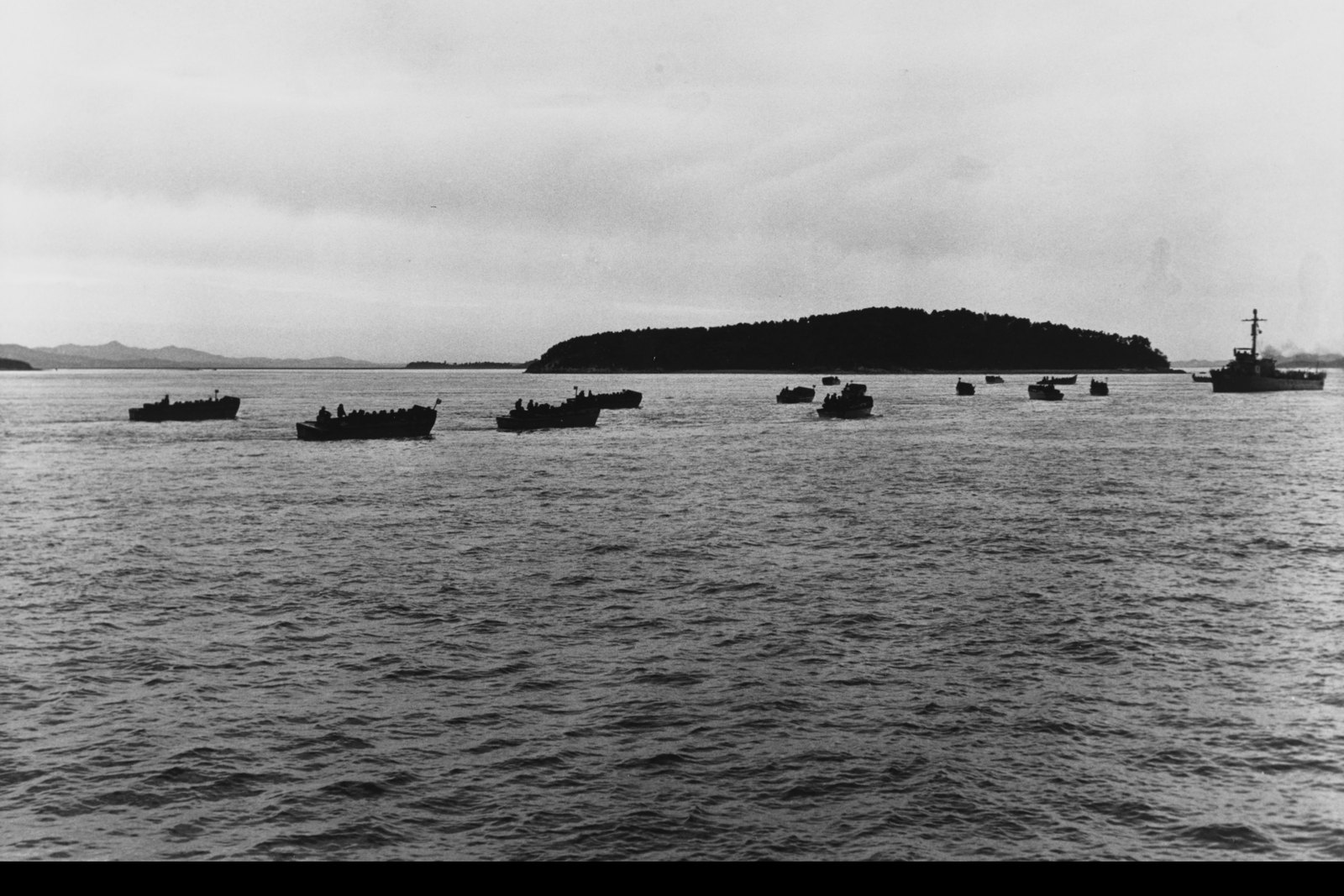 Black and white photograph of a gray sea under an overcast sky: a line of small landing craft packed with men heads toward a low wooded island, with a small warship on the right.