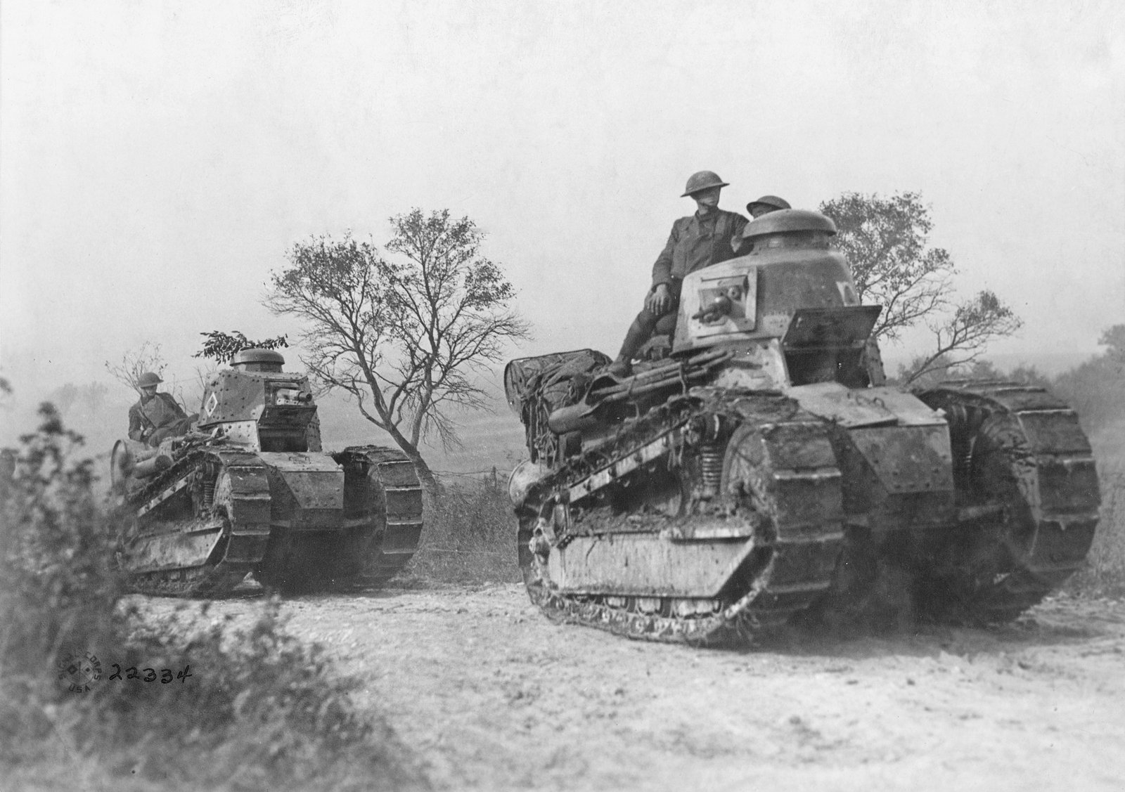 Black and white photograph of two small Renault FT tanks rolling along a dusty track past bare trees, with American soldiers in steel helmets riding on top of them.