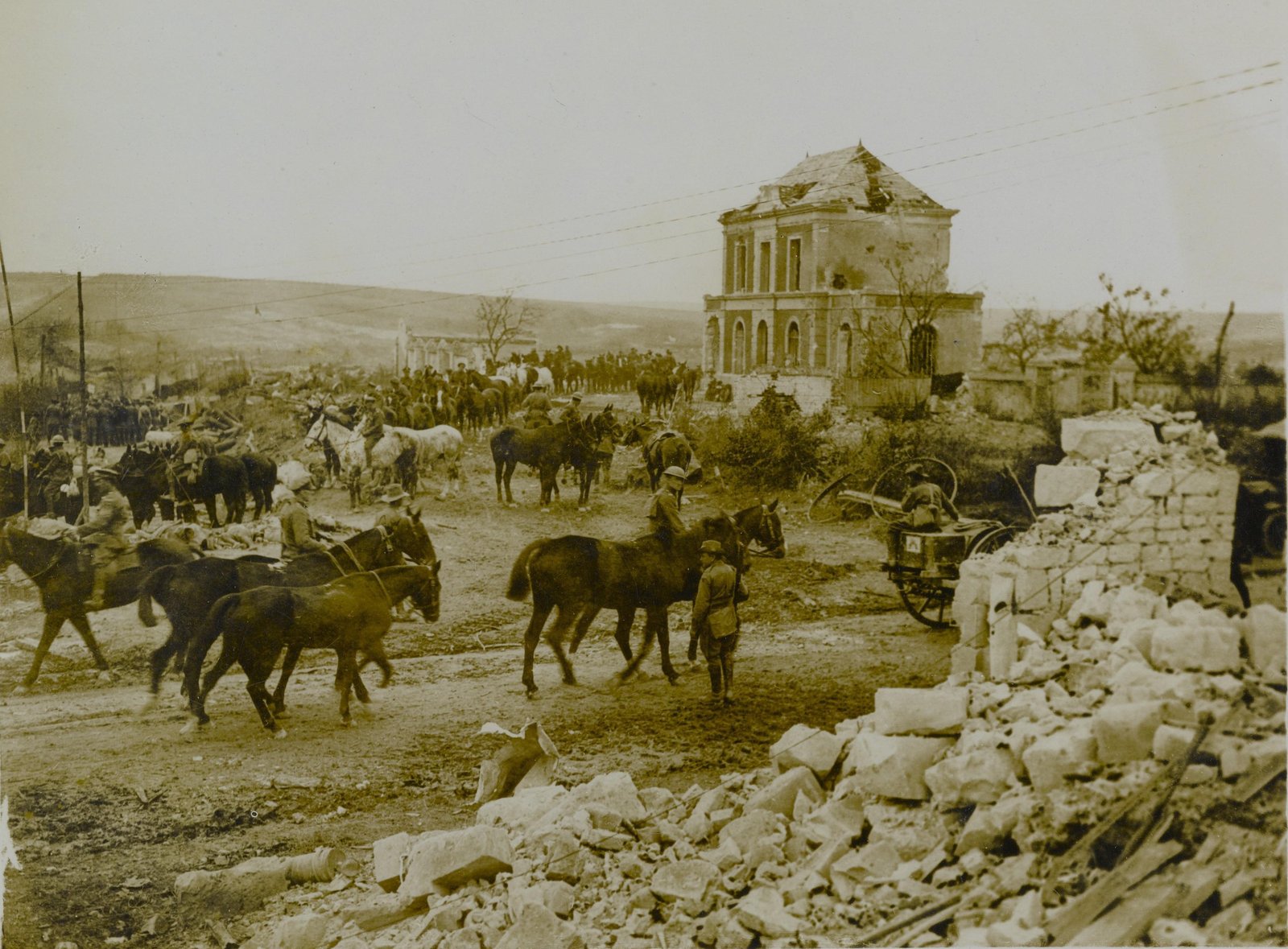 Sepia photograph of a ruined village street with mounted soldiers and horse lines among heaps of rubble, a shell-damaged two-storey building in the background and rolling bare hills beyond.